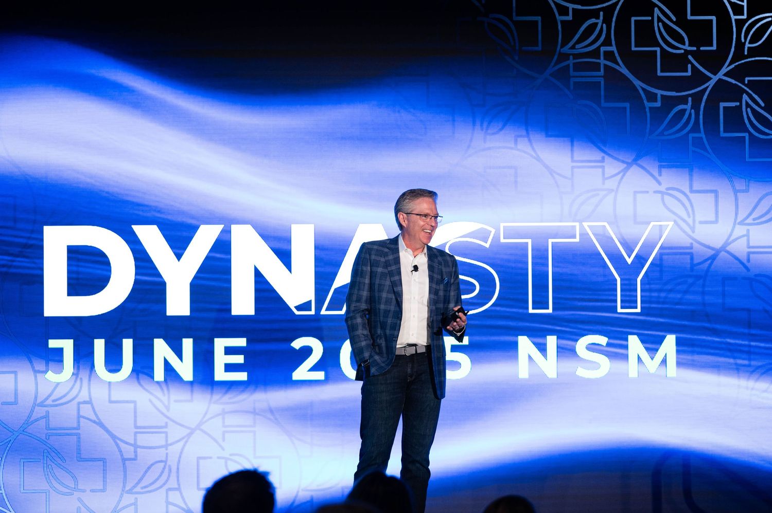 A presenter stands on stage in front of a large blue and white backdrop displaying the text 'DYNASTY JUNE' at a corporate event.