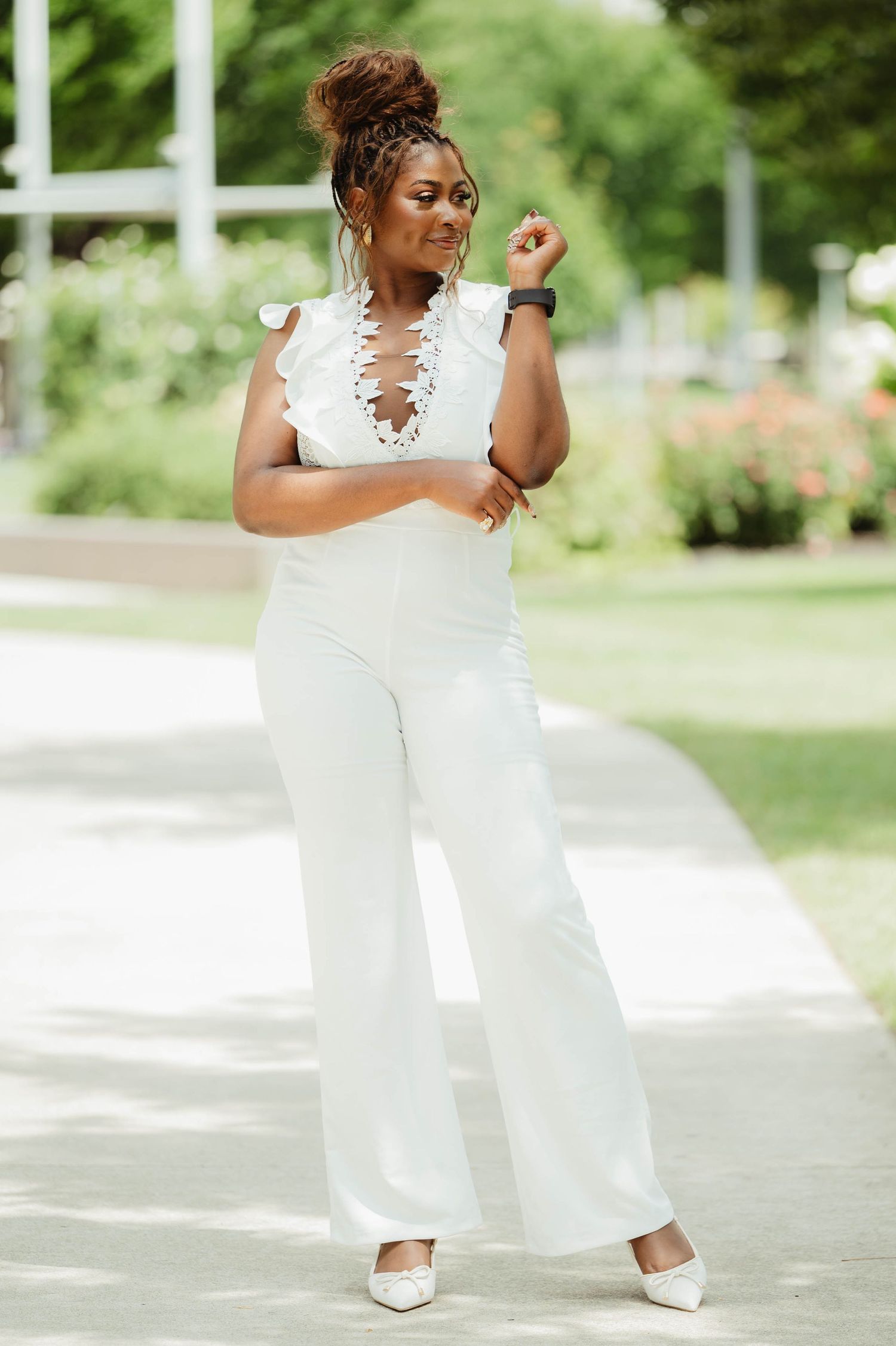 A person in an elegant white jumpsuit poses gracefully in a sunny outdoor garden setting.