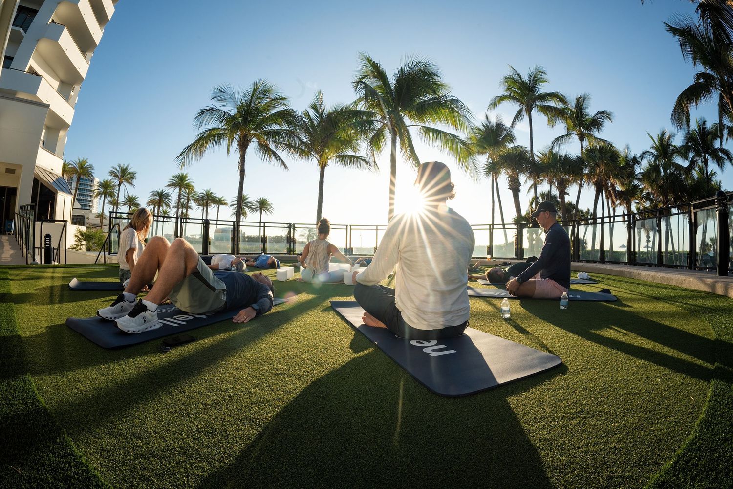 Group yoga class on green lawn with palm trees and sunny sky at a tropical resort.