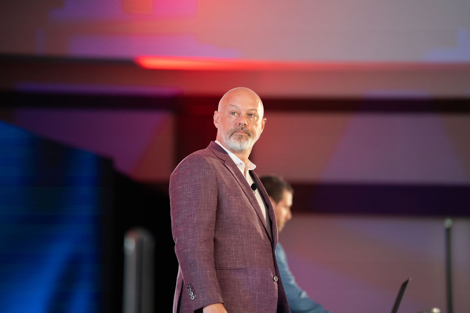Bald man with beard in maroon blazer presenting on stage at a professional conference event.