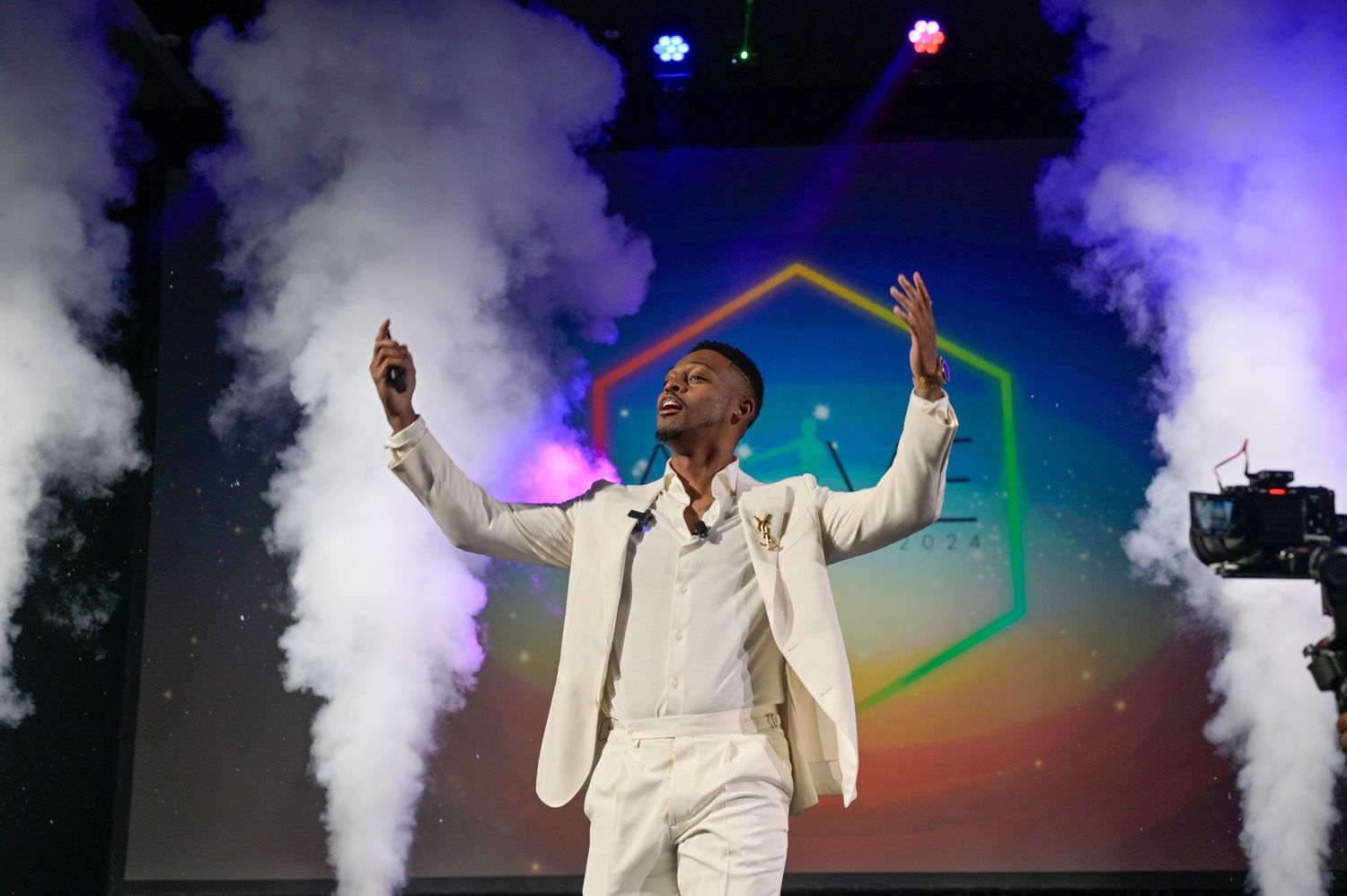 Performer in white suit on stage with colorful smoke effects and hexagon backdrop at live event.