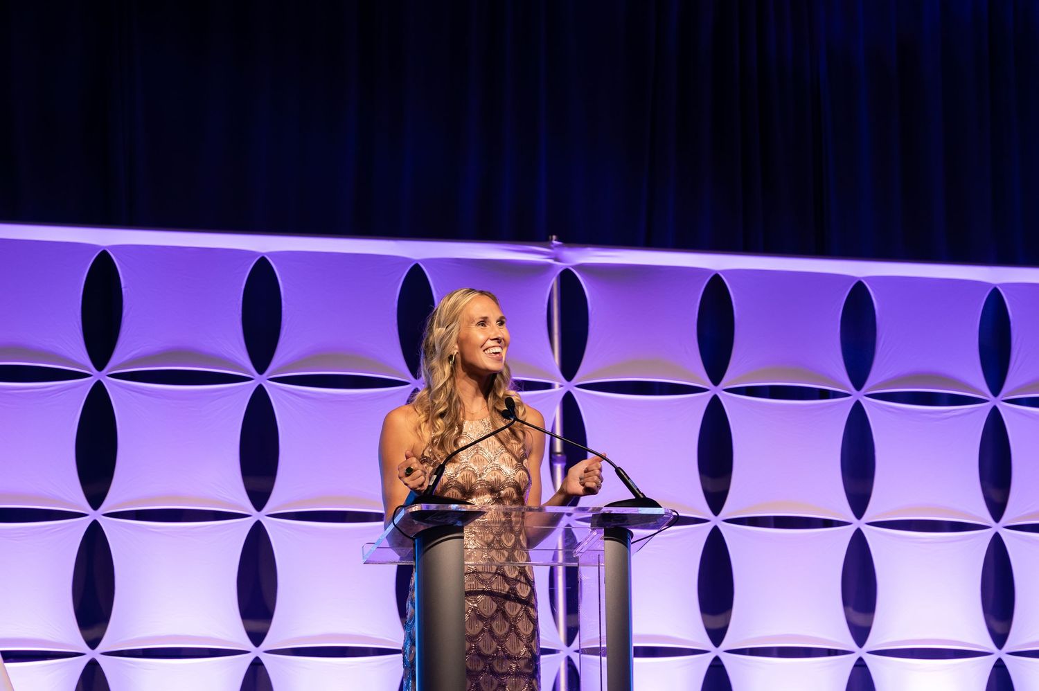 Woman speaking at podium on stage with purple geometric backdrop at formal awards ceremony.
