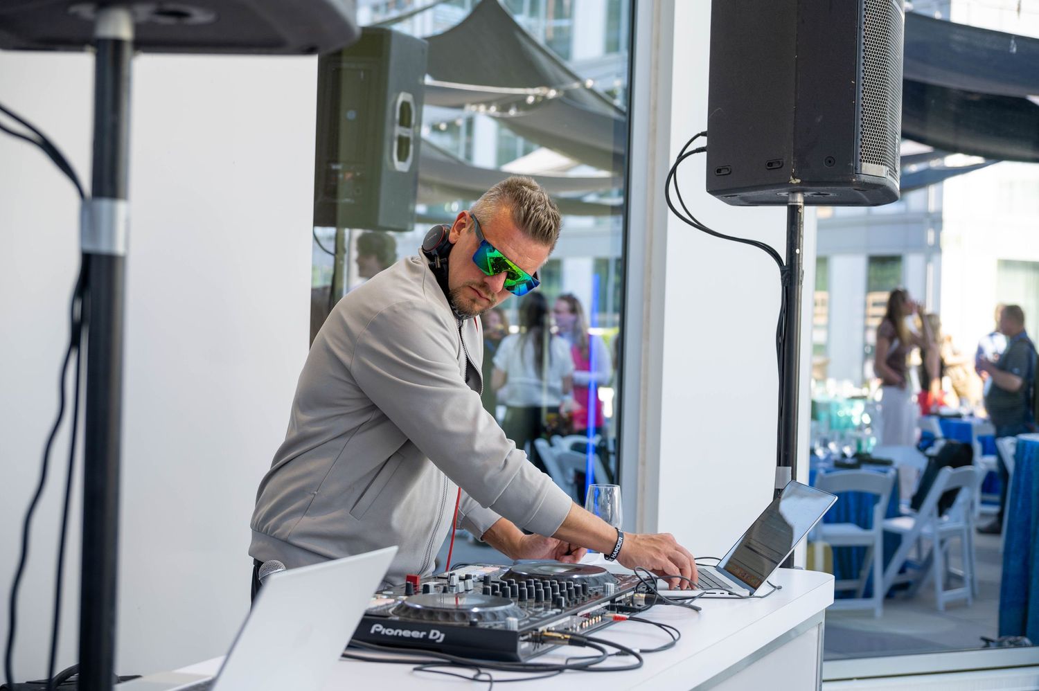 DJ wearing a gray jacket and orange headphones performs at an outdoor venue with mixing equipment.