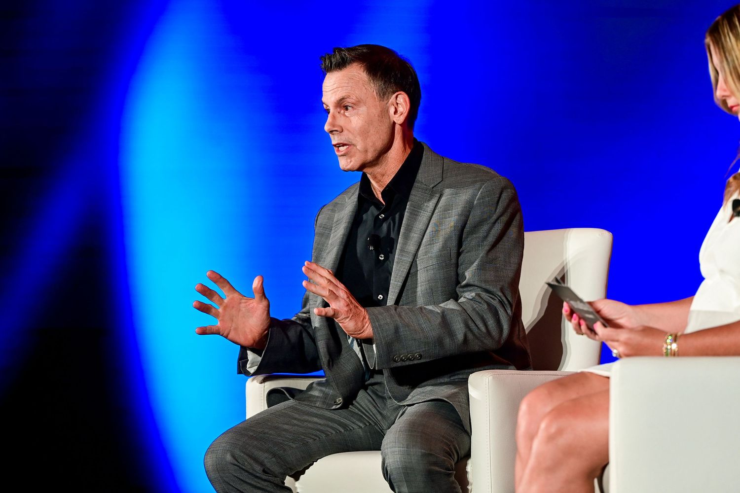 Man in gray blazer gestures while speaking on stage at a conference panel discussion.