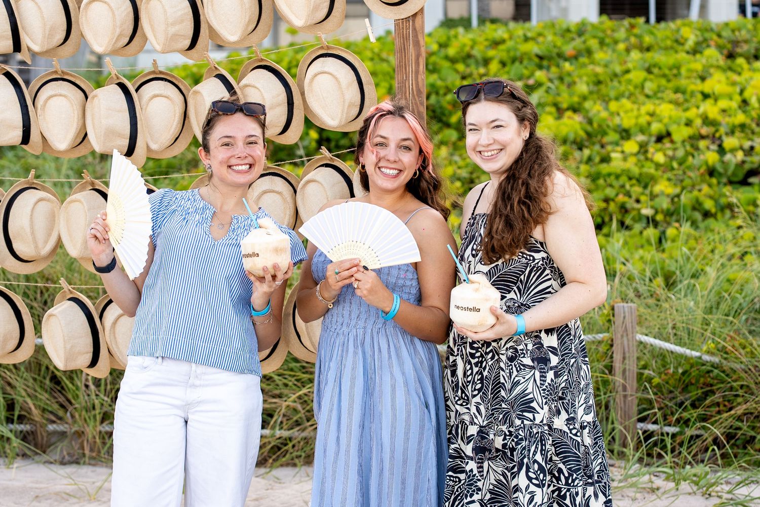 Three women smiling and holding fans near a wall of straw hats at a tropical beach destination.