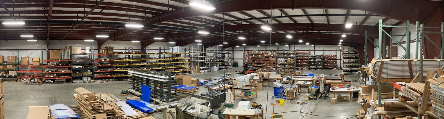 Panoramic view of a large industrial warehouse with shelving, materials, and equipment under bright overhead lighting.