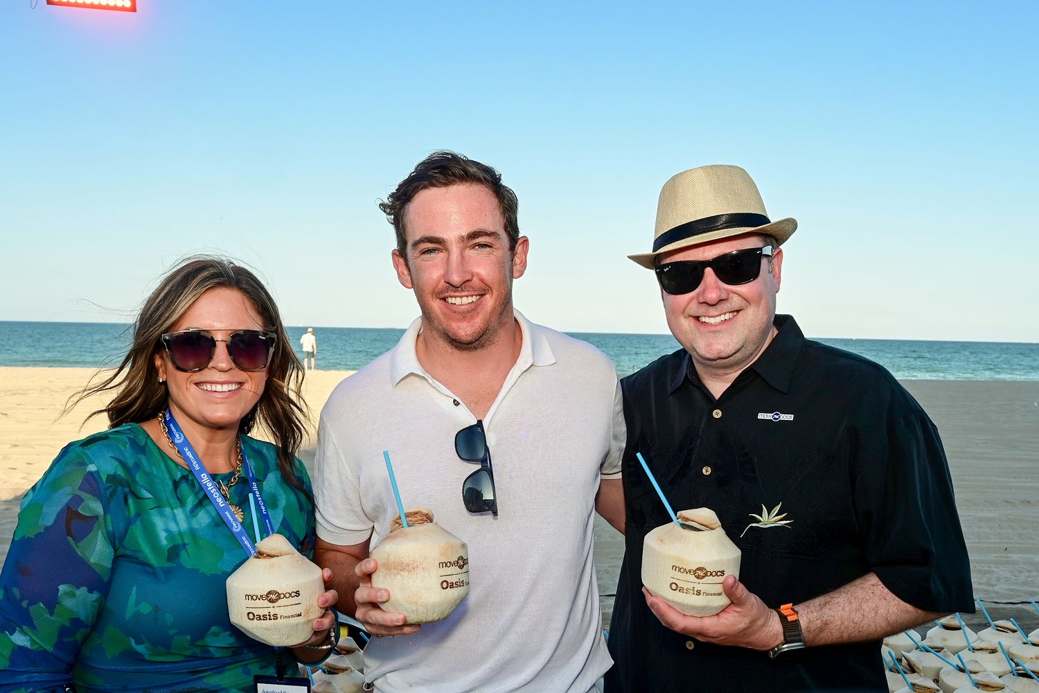Three guests smile and hold coconut drinks on a sunny beach at an outdoor social gathering.