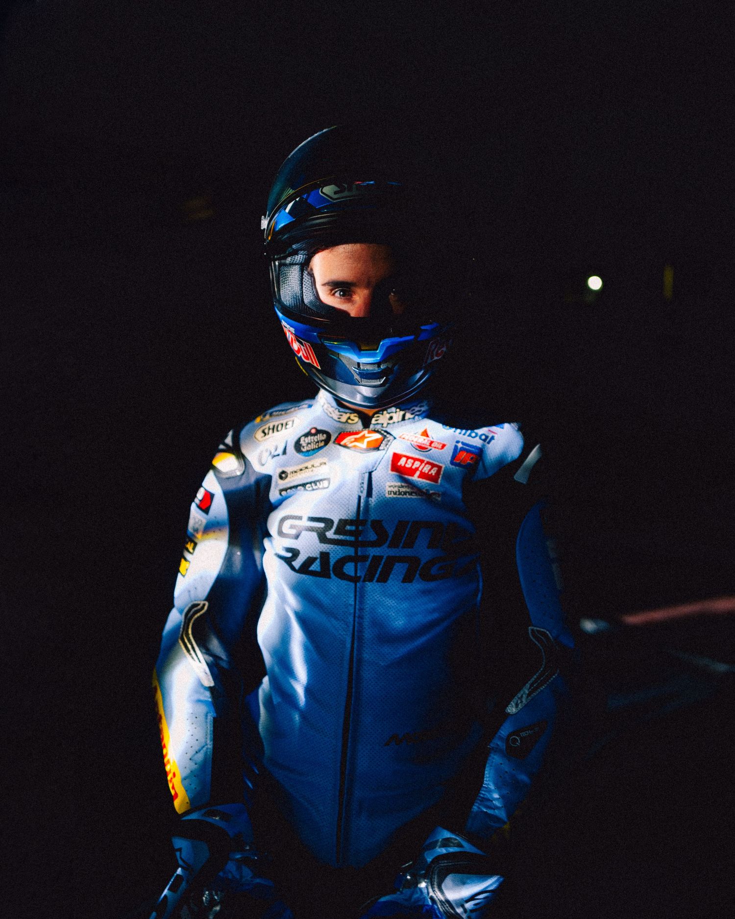 Racing motorcyclist in white and blue gear illuminated against dark background.