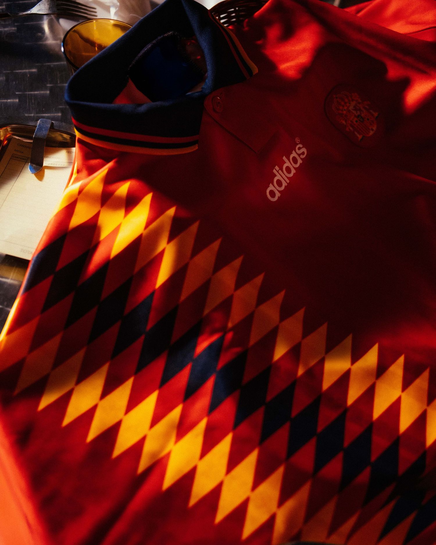 Adidas red and gold diamond pattern soccer jersey with navy collar displayed in warm sunlight.