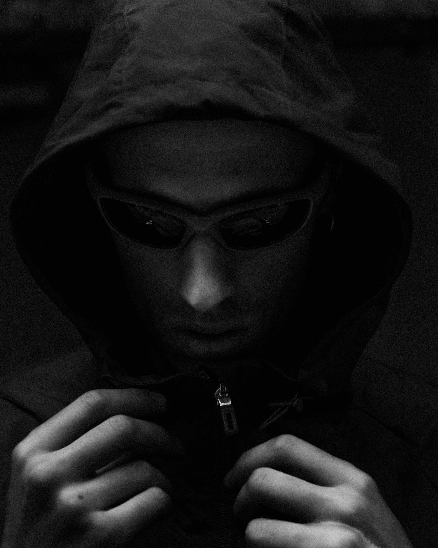 Black and white portrait of a person in a dark hoodie wearing sleek sunglasses, looking down mysteriously.