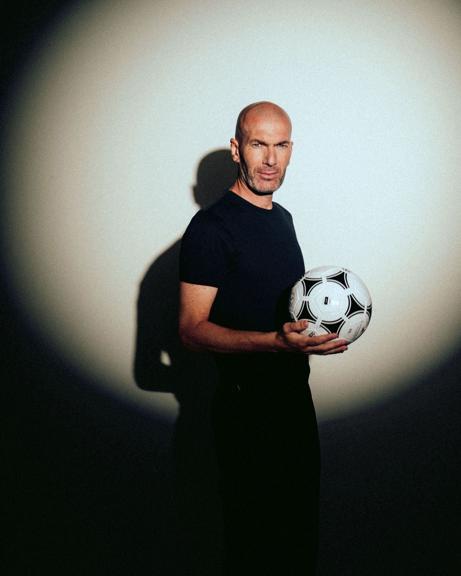 A figure in black clothing poses with a soccer ball against a white backdrop with dramatic lighting and shadows.