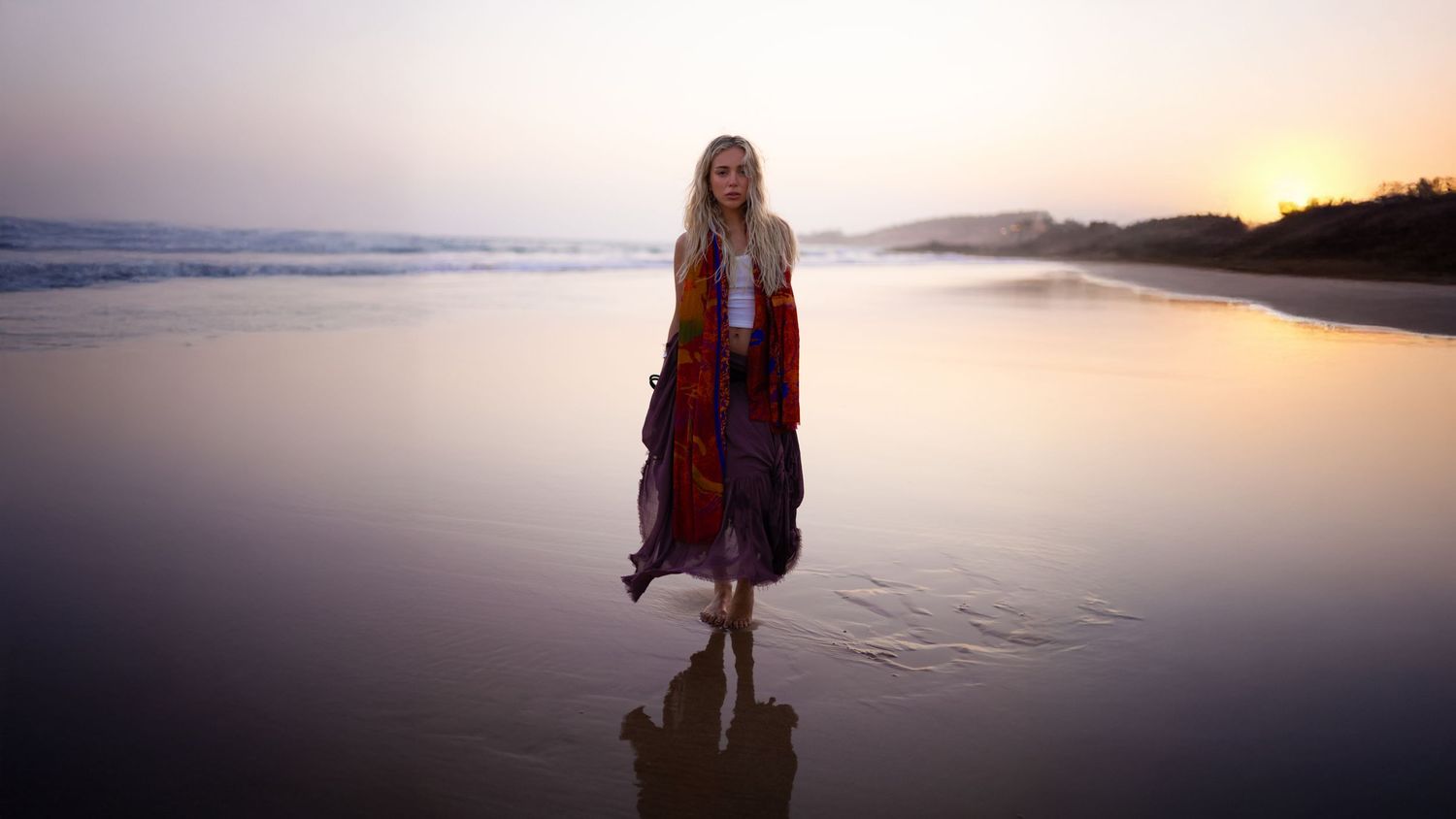 A silhouetted figure walks along a tranquil beach at sunset with calm ocean waters and golden sky in the background.