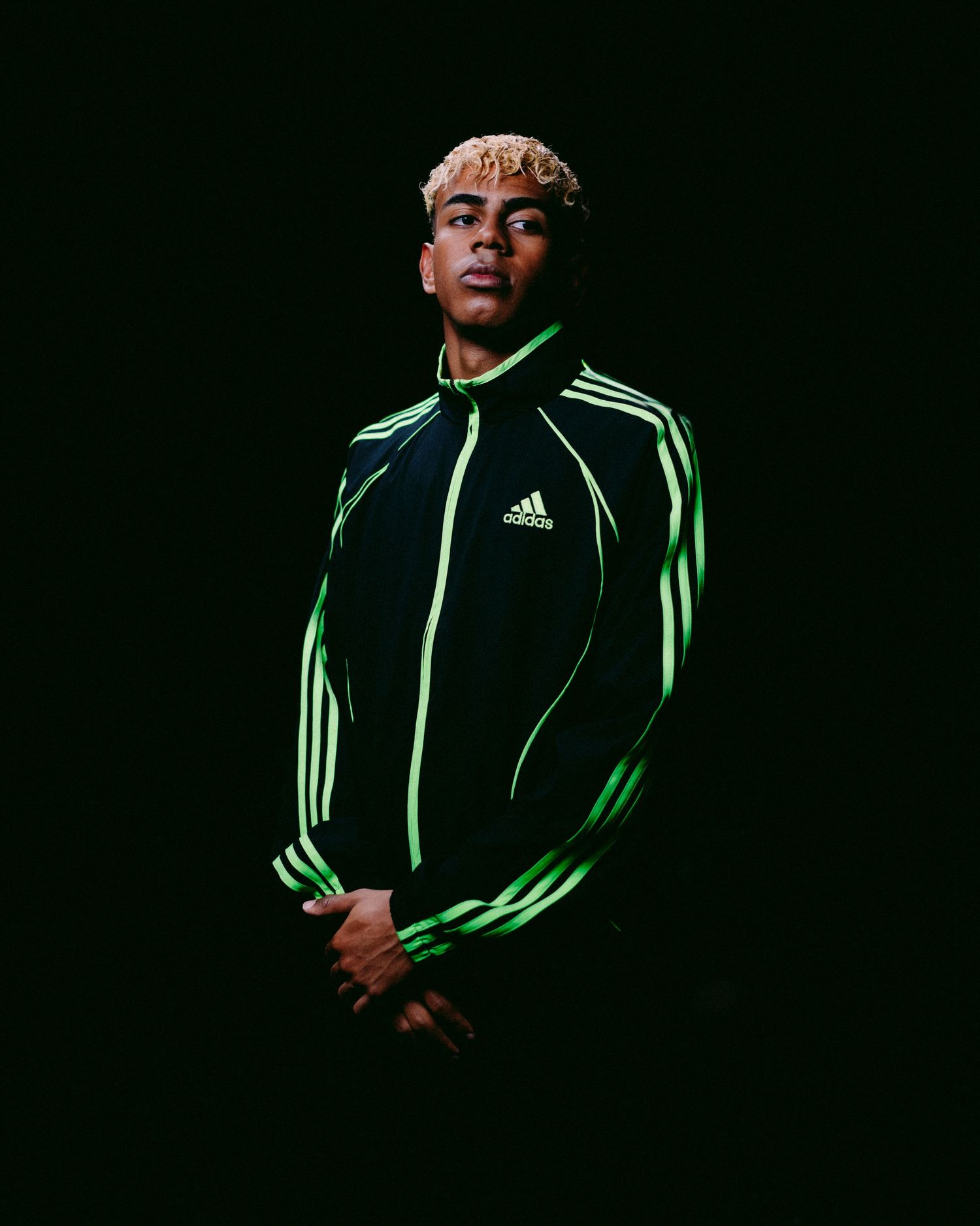 Dark dramatic shots of someone wearing a green-striped Adidas jacket against black background.
