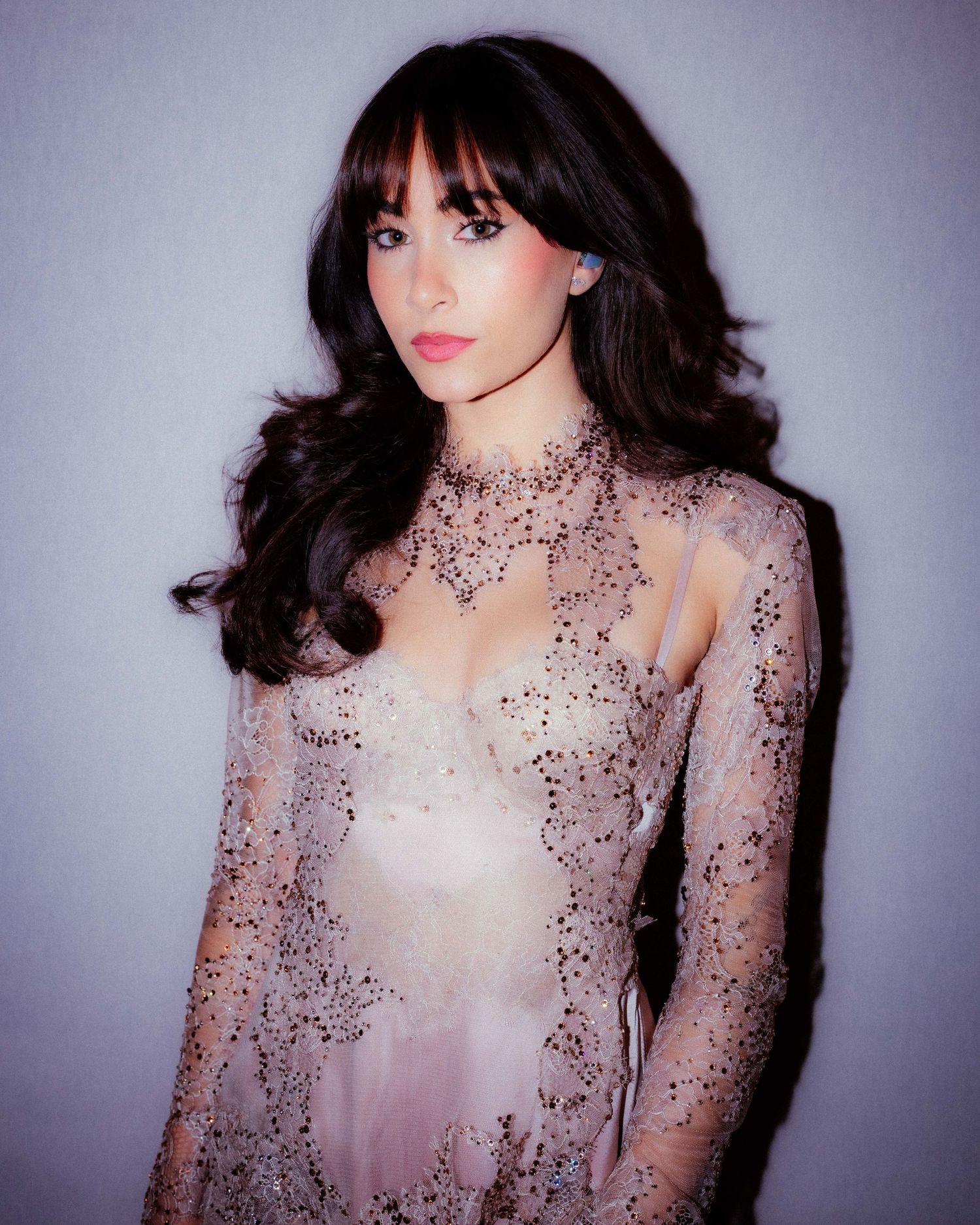 Young woman with dark wavy hair and bangs wearing an elegant sheer lace embellished gown posing against a light background.