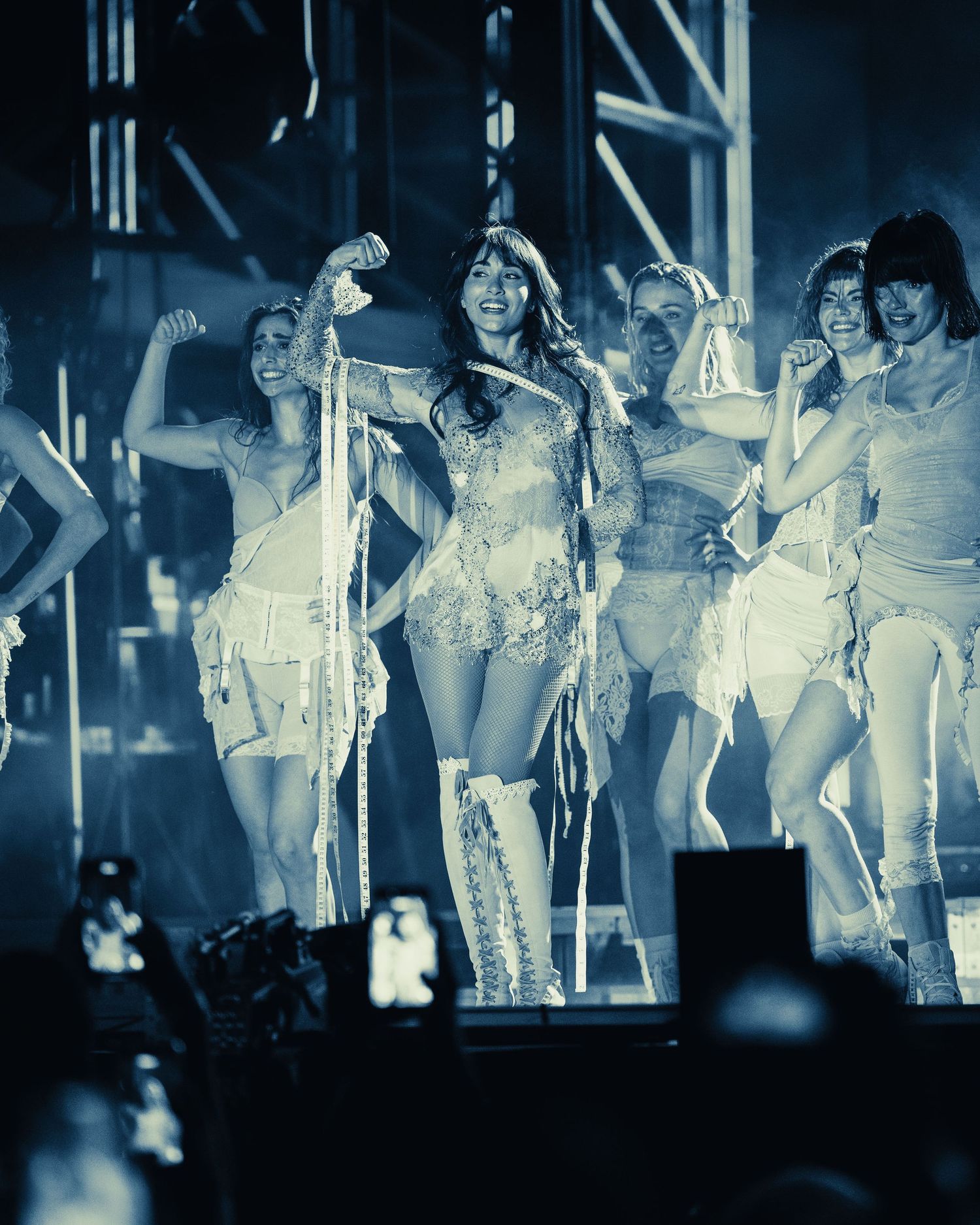Performer in ornate costume on stage with dancers in blue-toned concert photo.