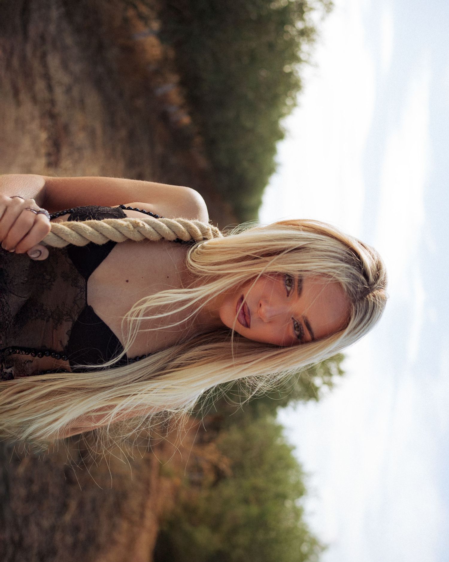A woman with long blonde hair poses outdoors in a wooded area with soft natural lighting.