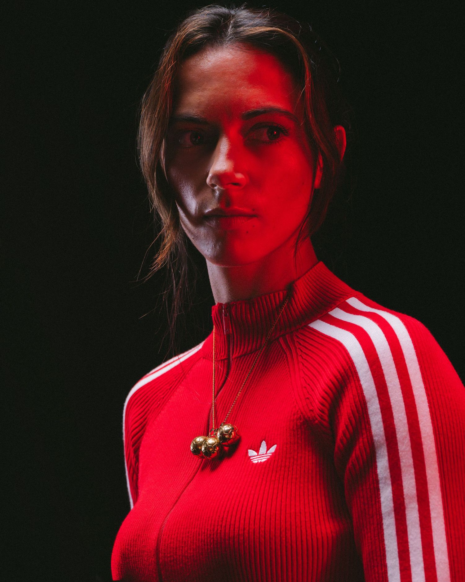 Dramatic portrait in red lighting showing someone wearing a red Adidas track jacket with classic three stripes against dark background.