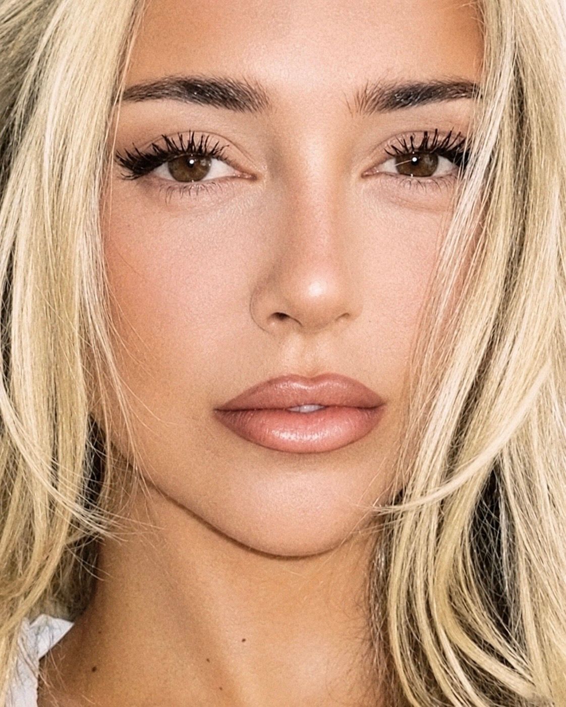 Close-up portrait of a young woman with blonde hair, brown eyes, long lashes, and glossy nude lips.