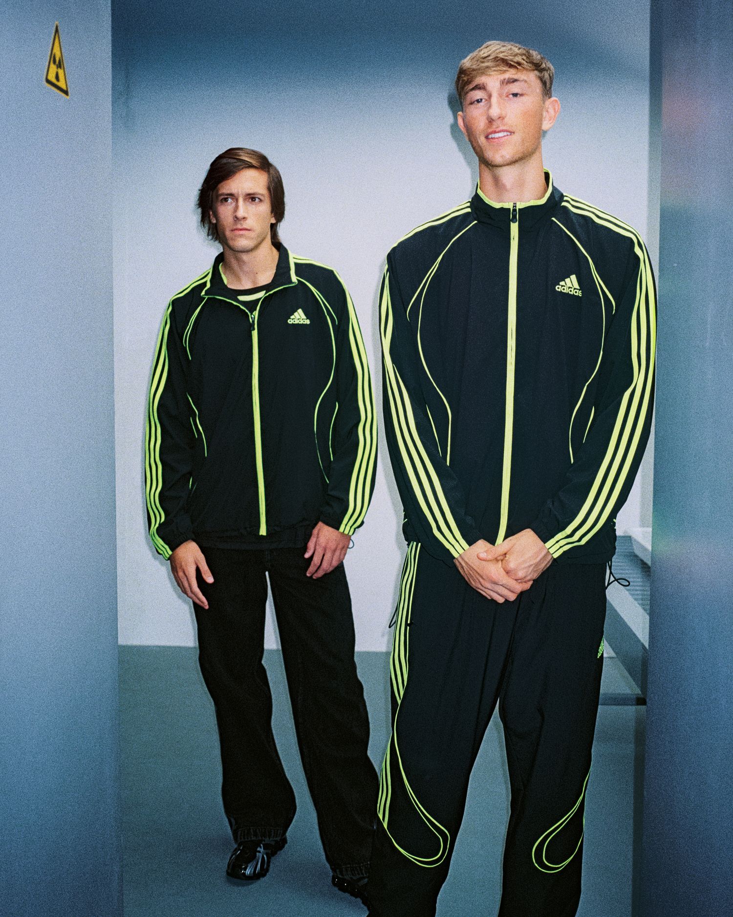 Two people pose together wearing matching black Adidas tracksuits with neon yellow stripes in a studio setting.