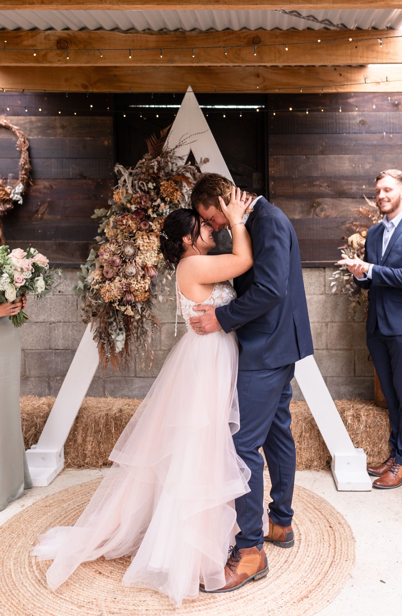 Intimate wedding moment in front of a triangular arch with dried flowers and wooden walls.