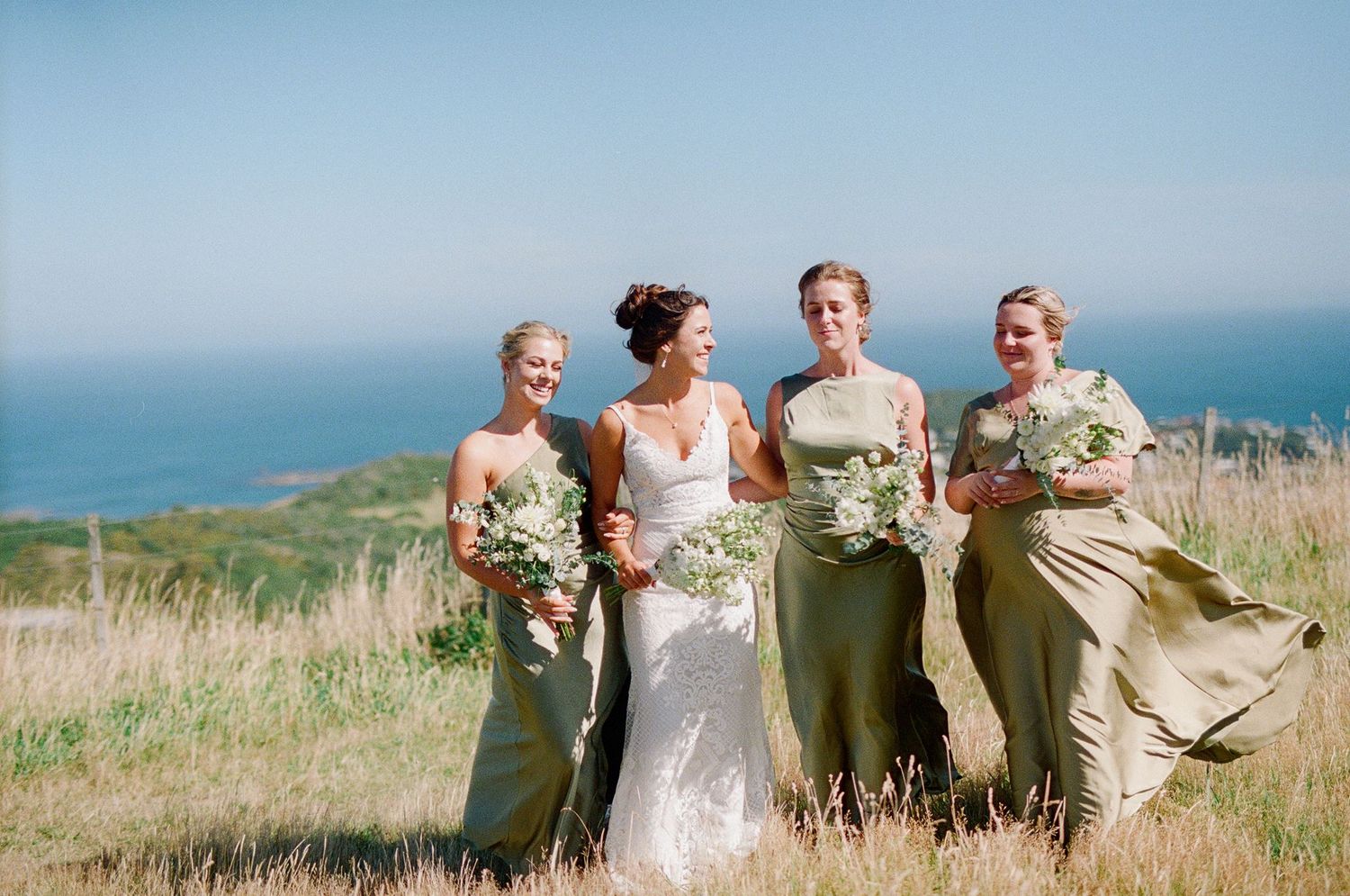Bride in white gown with three bridesmaids in sage green dresses holding bouquets on a scenic hillside overlooking the ocean.