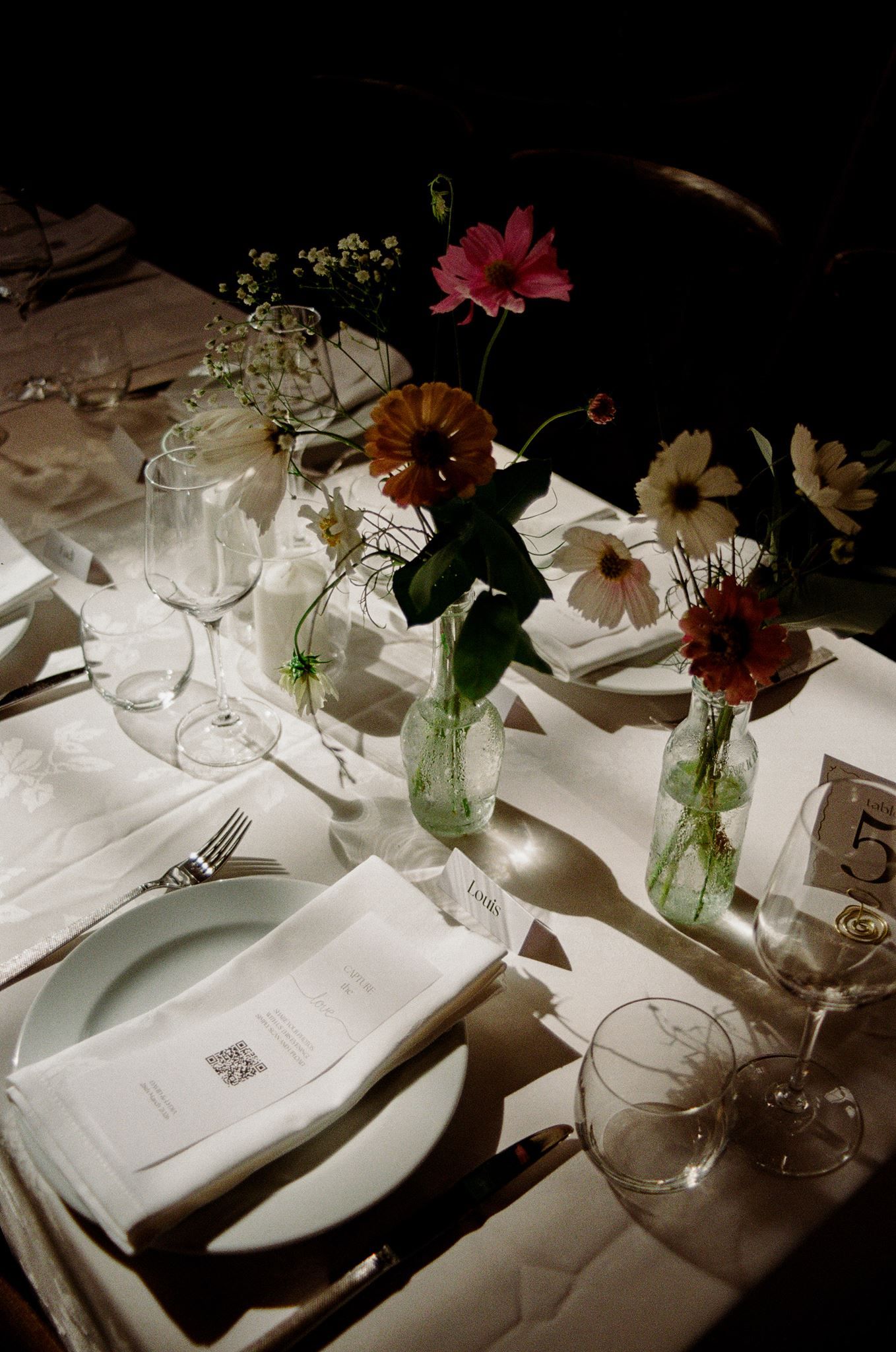 Elegant wedding table setting with colorful gerbera daisies in glass vases, white linens, crystal glasses, and a menu card.