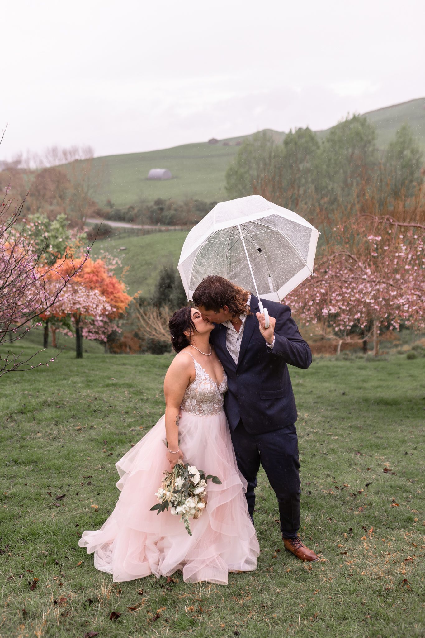 A romantic wedding photo of a couple with a clear umbrella in a garden with spring blossoms.