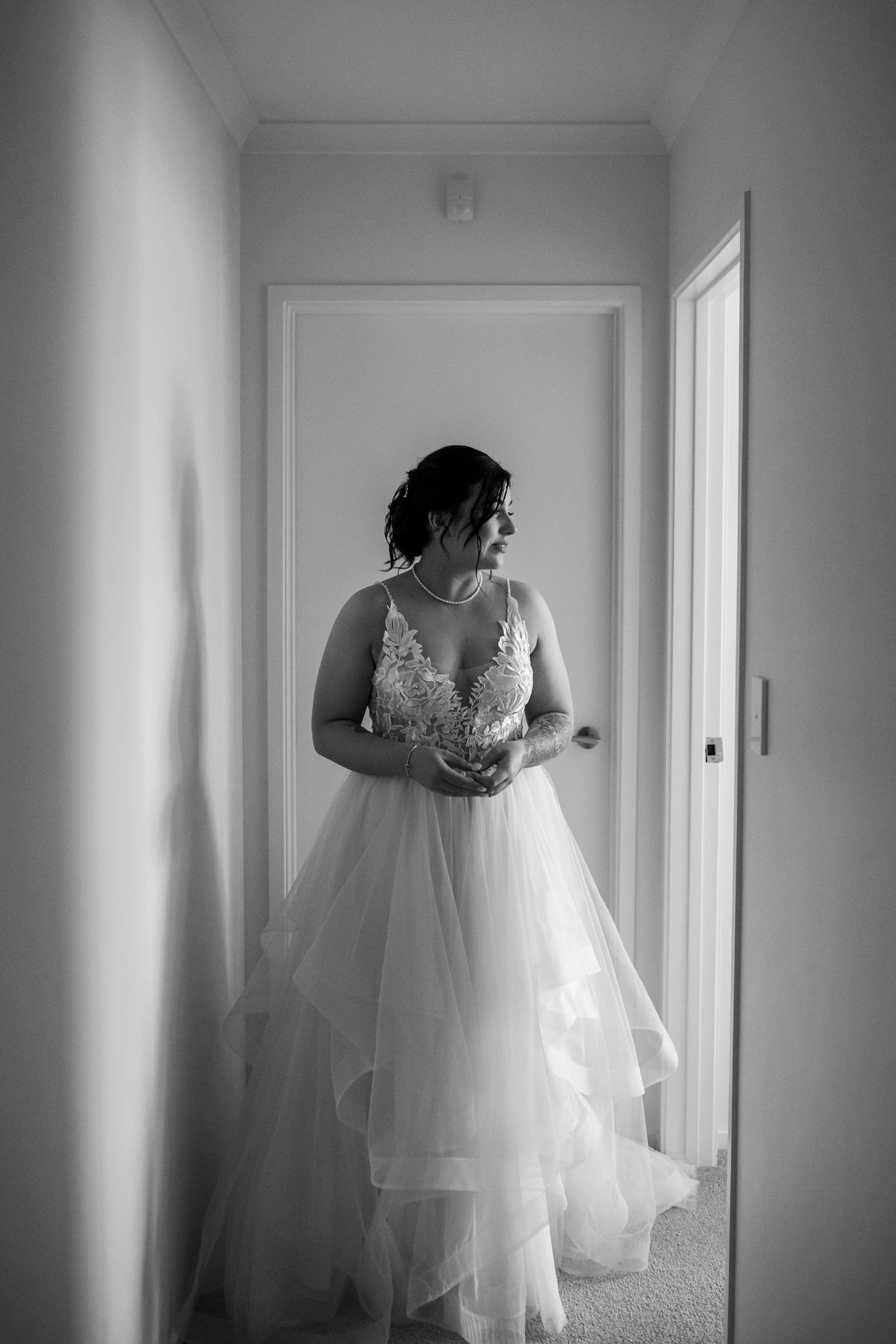 A black and white photo of a bride in an elegant lace and tulle wedding gown standing in a hallway.