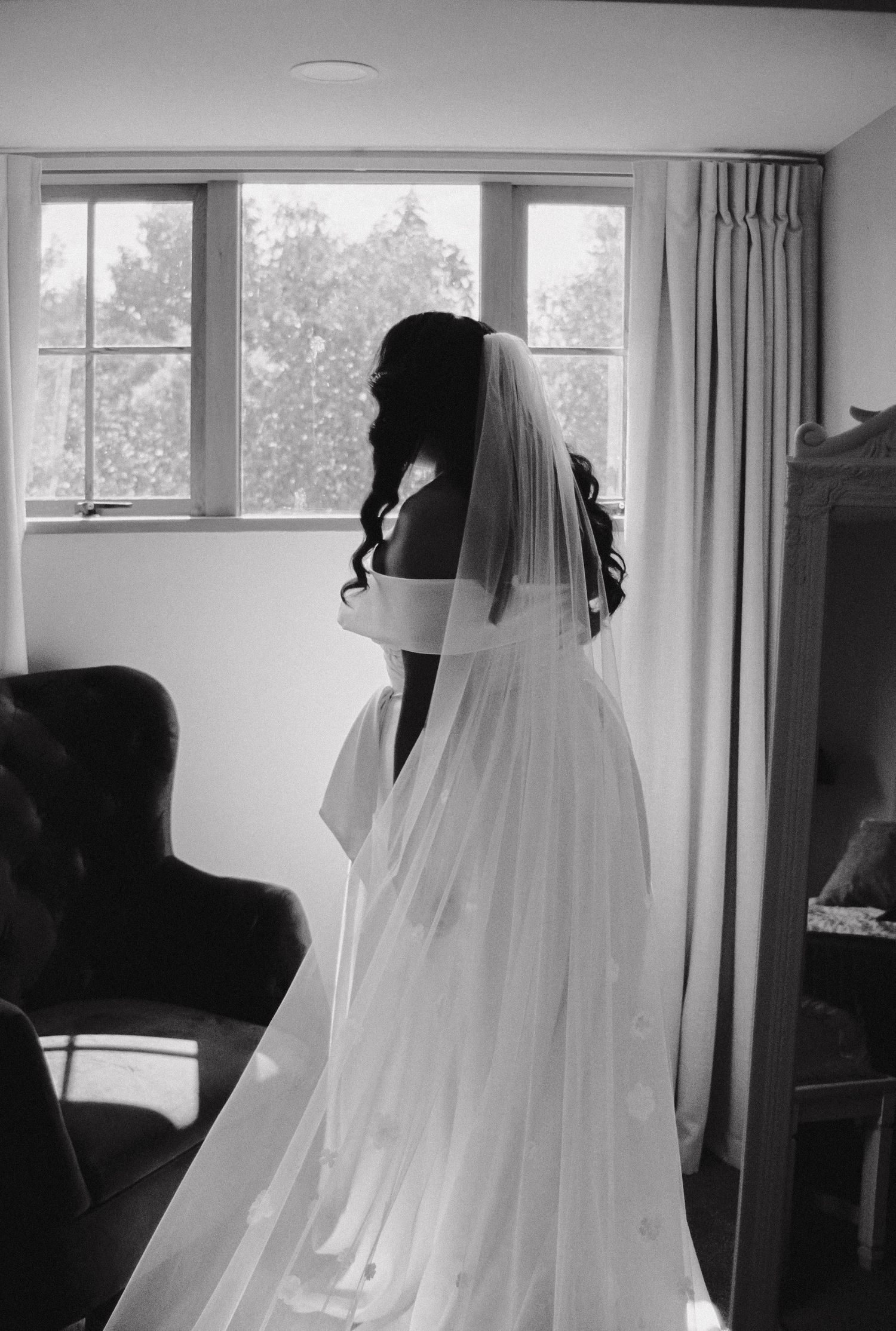 Black and white photo of a bride in a flowing white gown and veil gazing out of a window on her wedding day.