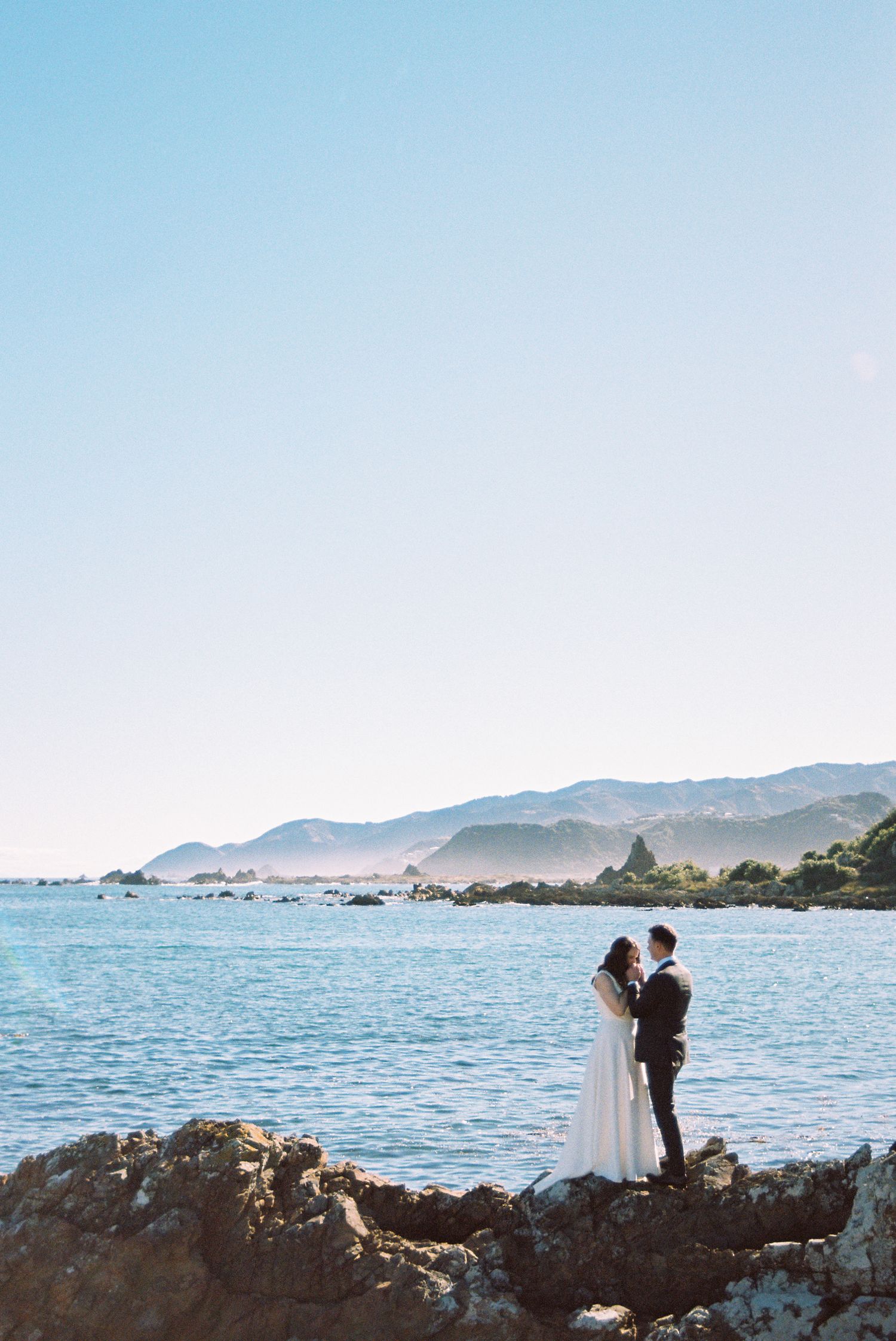 A scenic coastal wedding photo session with a couple embracing on rocky cliffs overlooking the ocean and mountains.