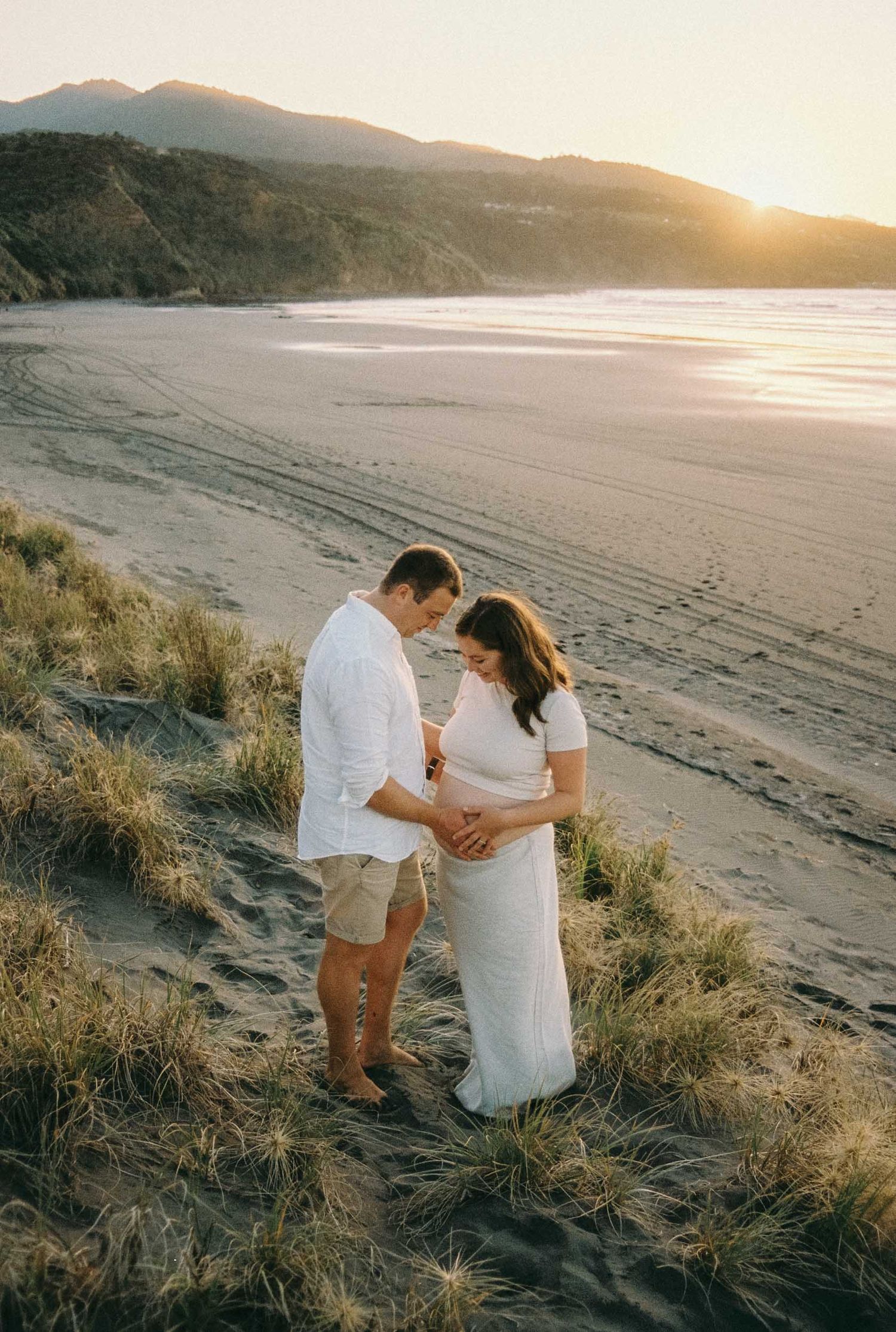 A couple embraces on a sandy beach at sunset with mountains and coastline in the background. Maternity photoshoot Wellington and Waikato