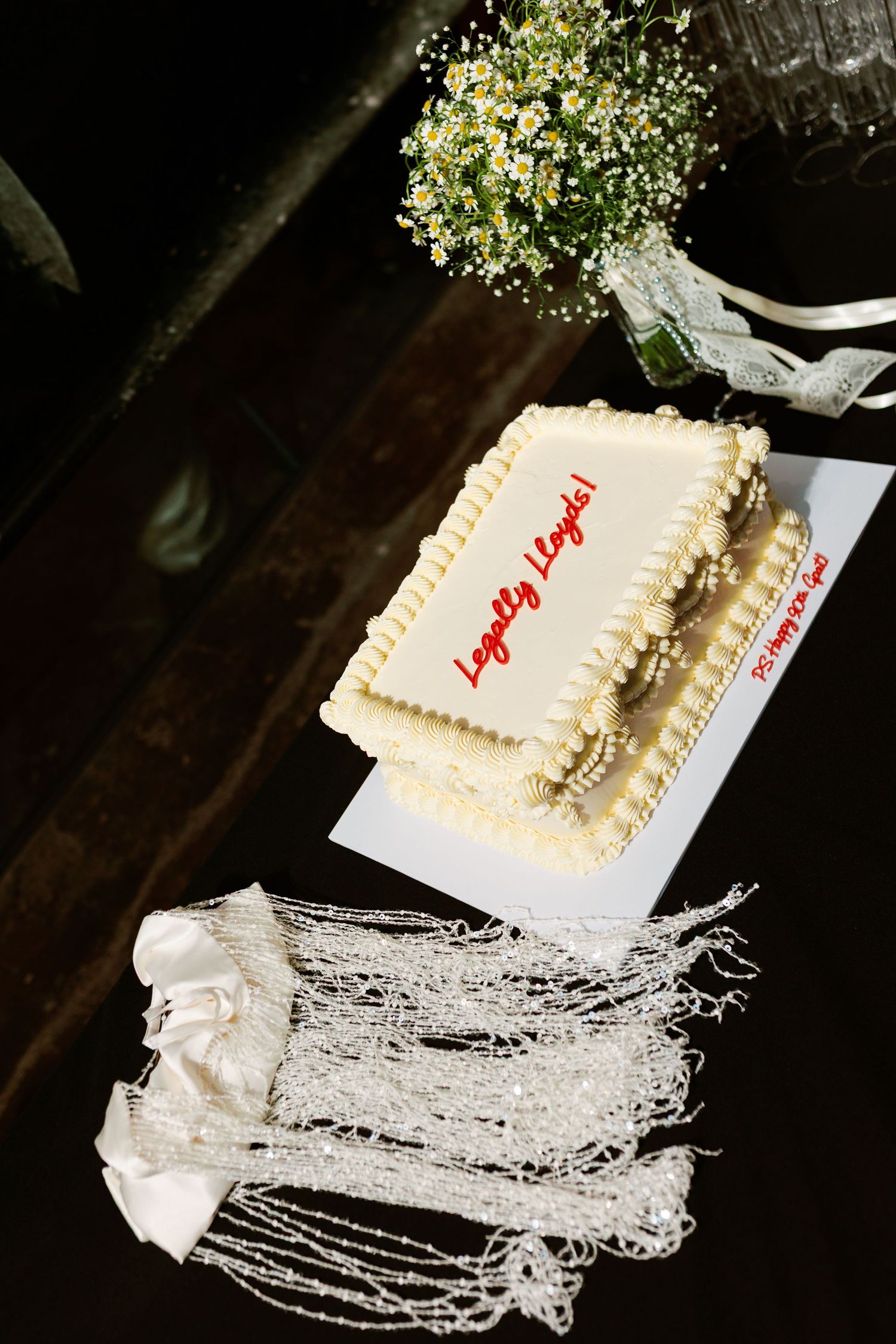 White square wedding cake with red “Legally Legally” text, baby's breath bouquet, and beaded accessories on dark table.