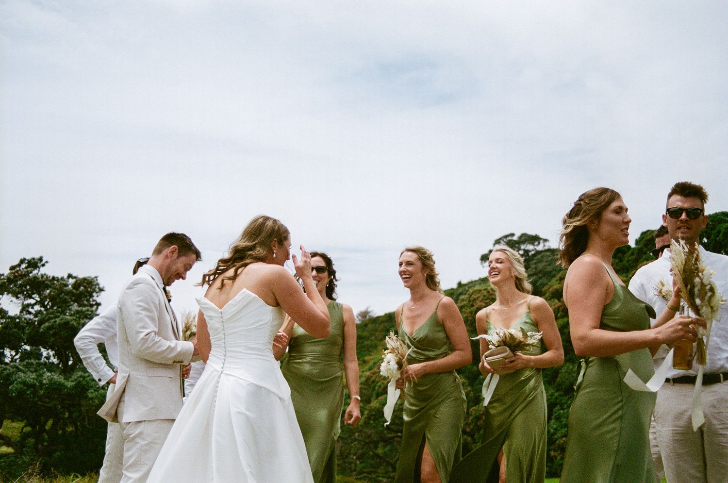 Wedding party members in olive green dresses and white suits pose together outdoors on a grassy hill.