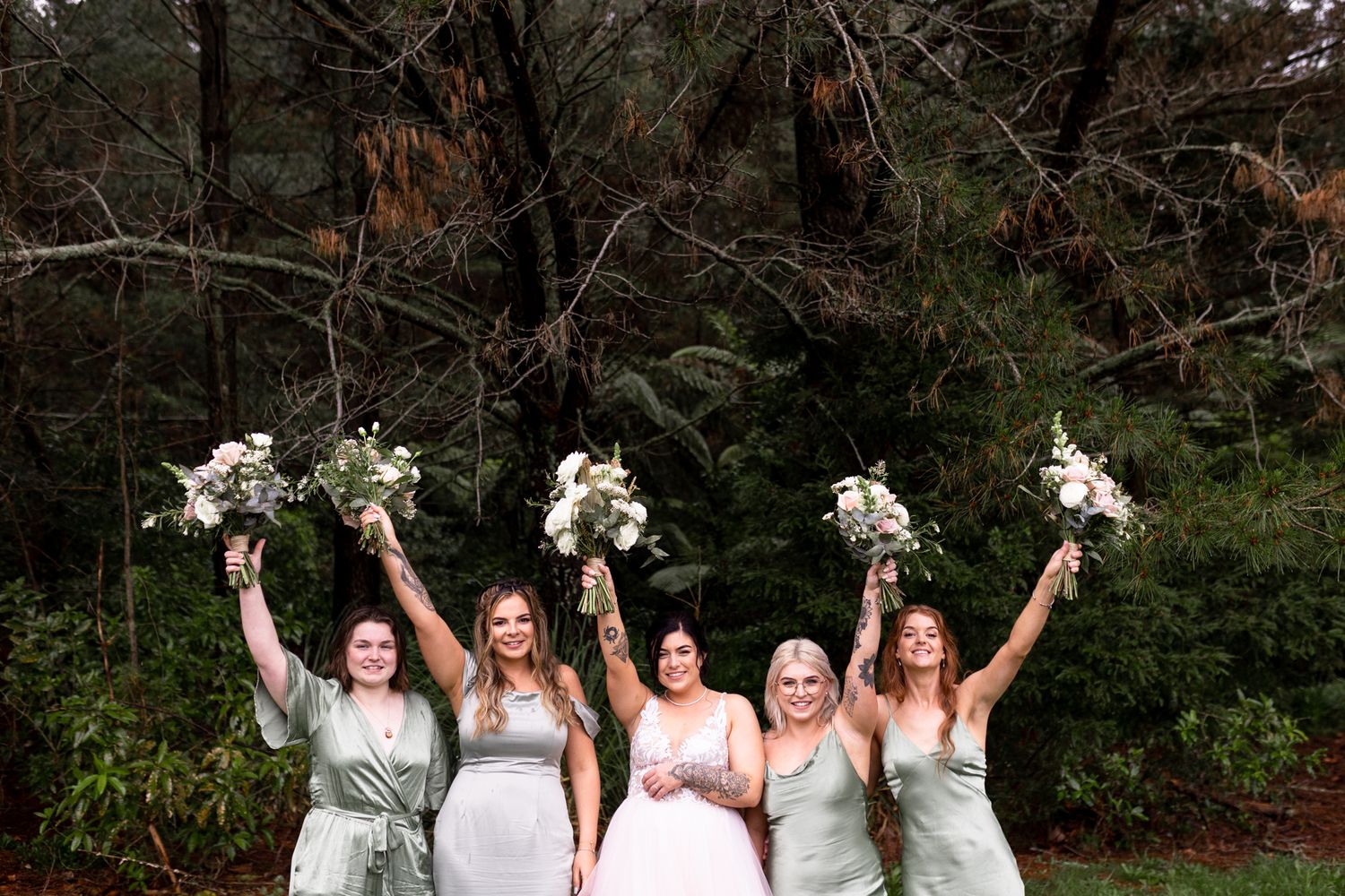Bridesmaids in sage green dresses hold bouquets high while posing against a forest backdrop.