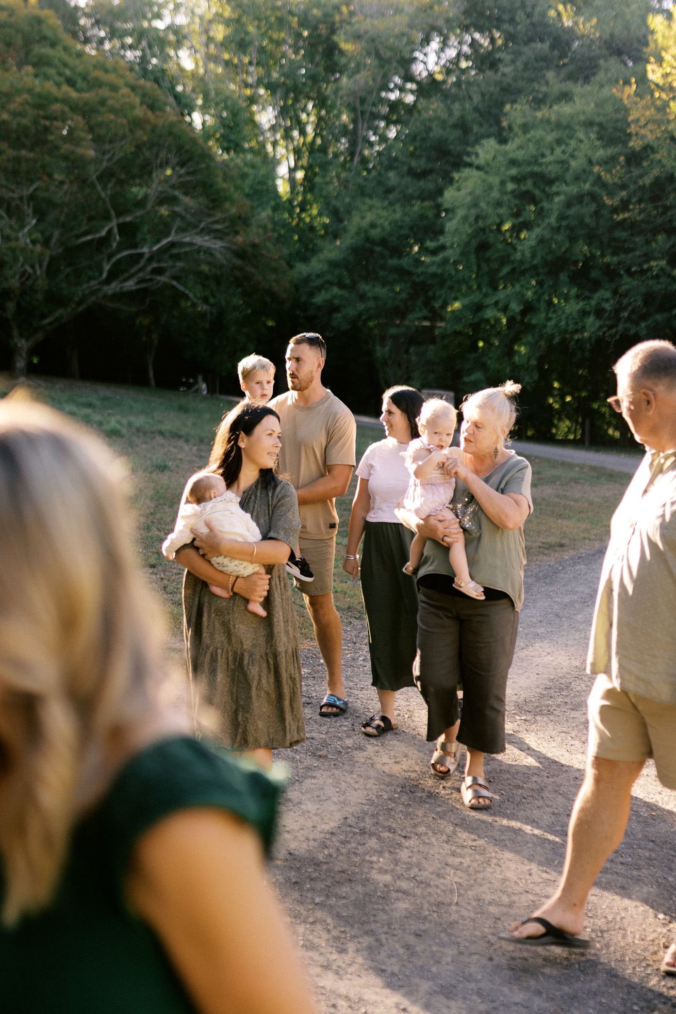 People gather together on a dirt path surrounded by trees on a sunny evening, dressed in neutral earth-toned attire.