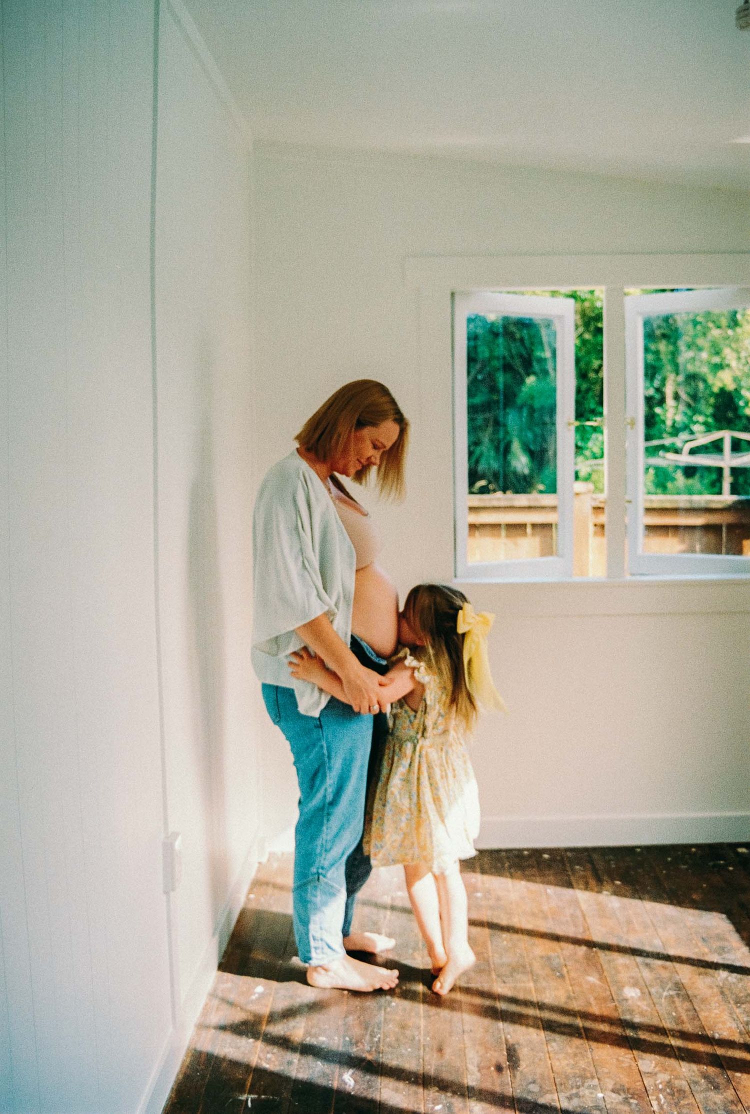 A tender moment between parent and child holding hands in an empty room with sunlit windows and wooden flooring. Intimate family photo