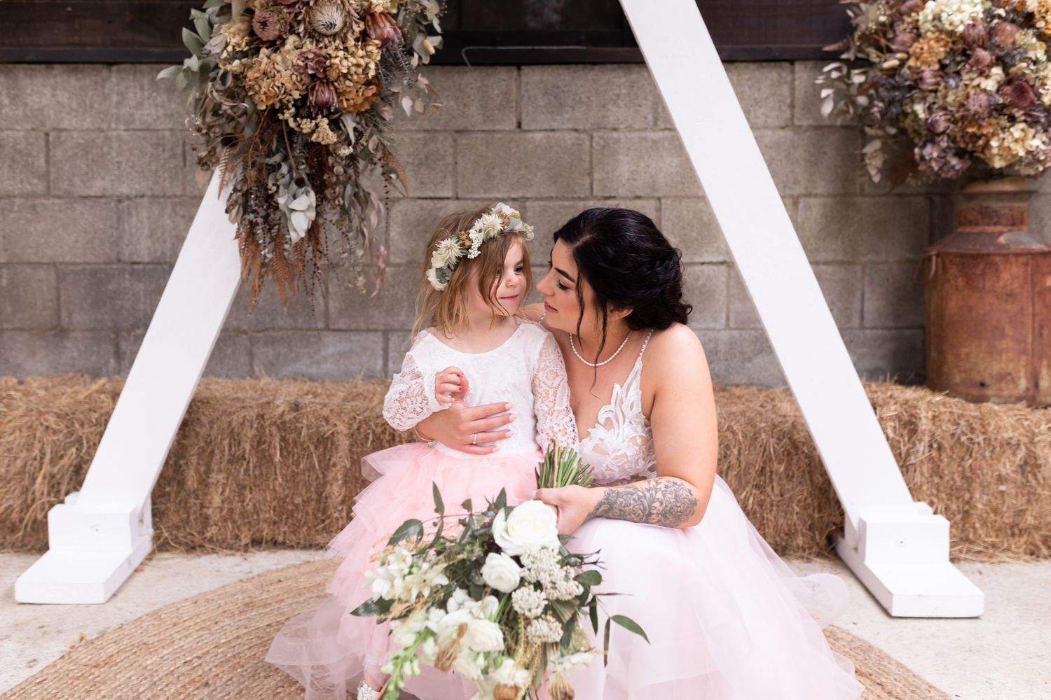 Two brides in pink dresses share a moment on hay bales under rustic floral decorations.