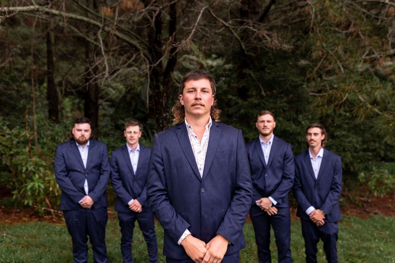 Groomsmen in navy suits pose for formal portrait in front of evergreen trees.