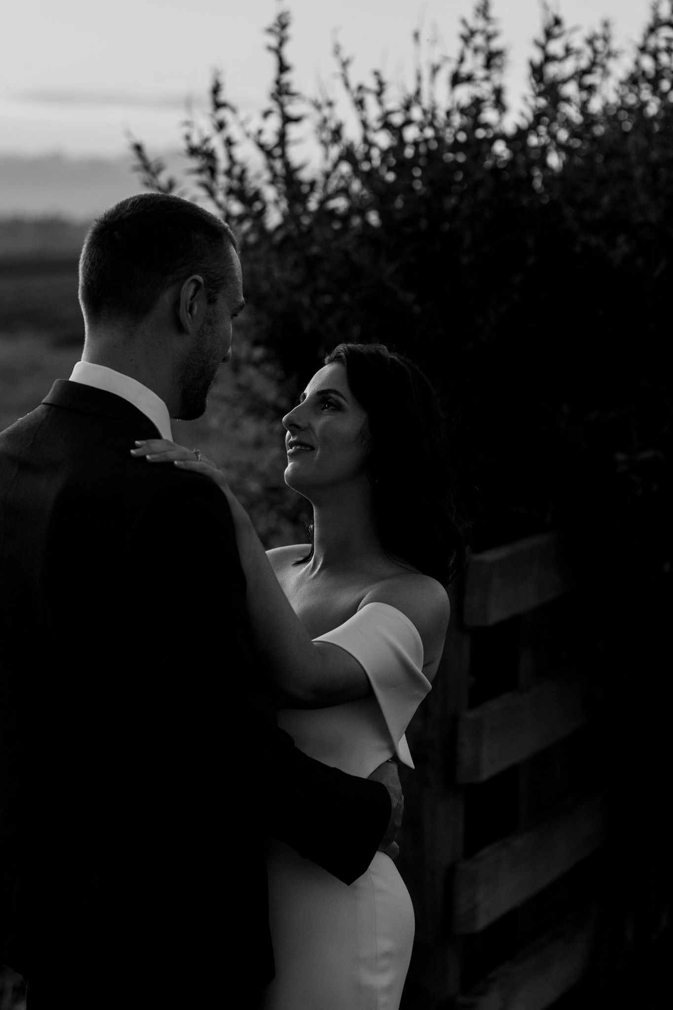 Romantic black and white portrait of newlyweds sharing an intimate moment against foliage at sunset.