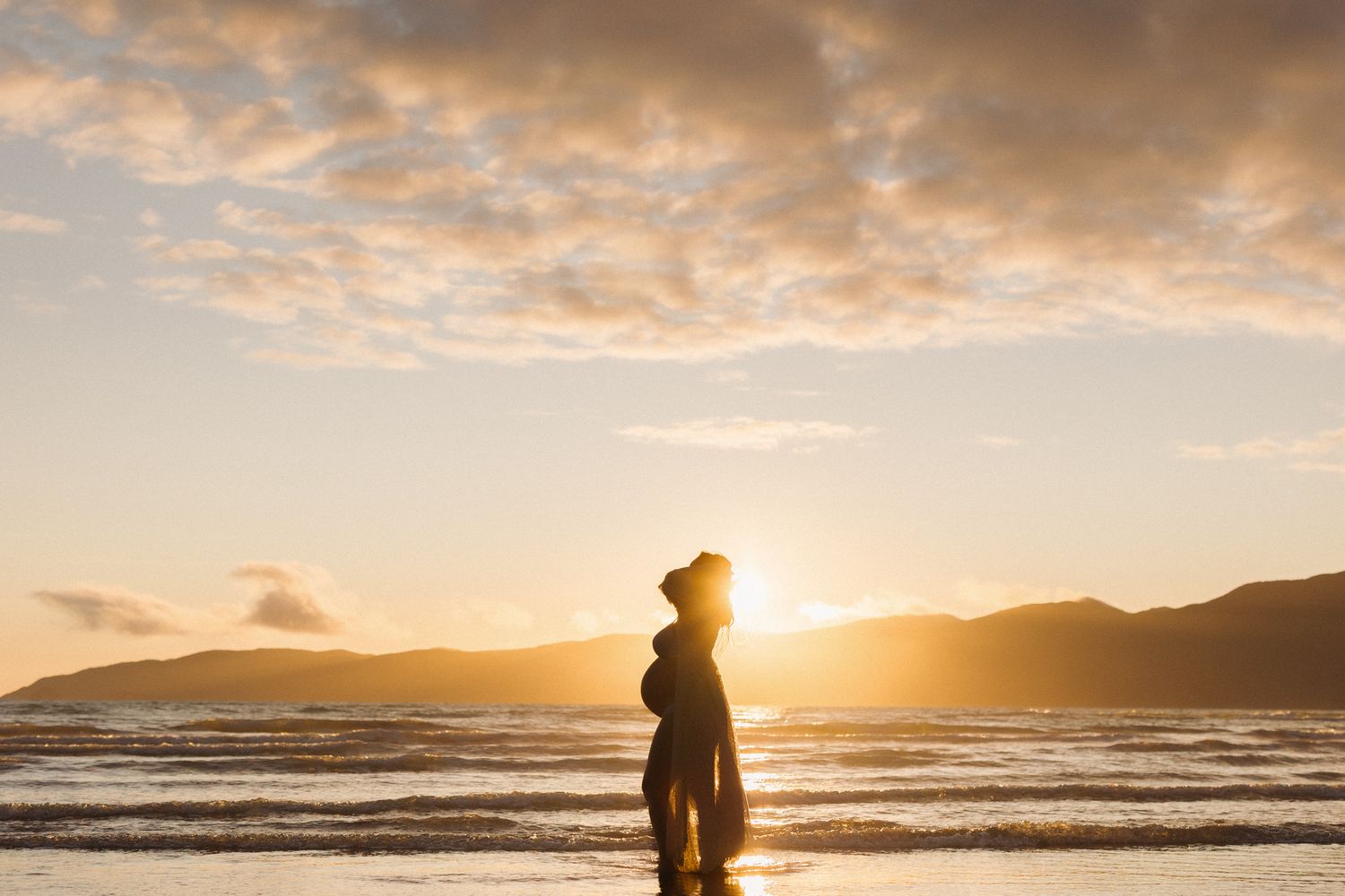 A silhouetted figure stands on a sandy beach during a dramatic golden sunset over the ocean waves.
