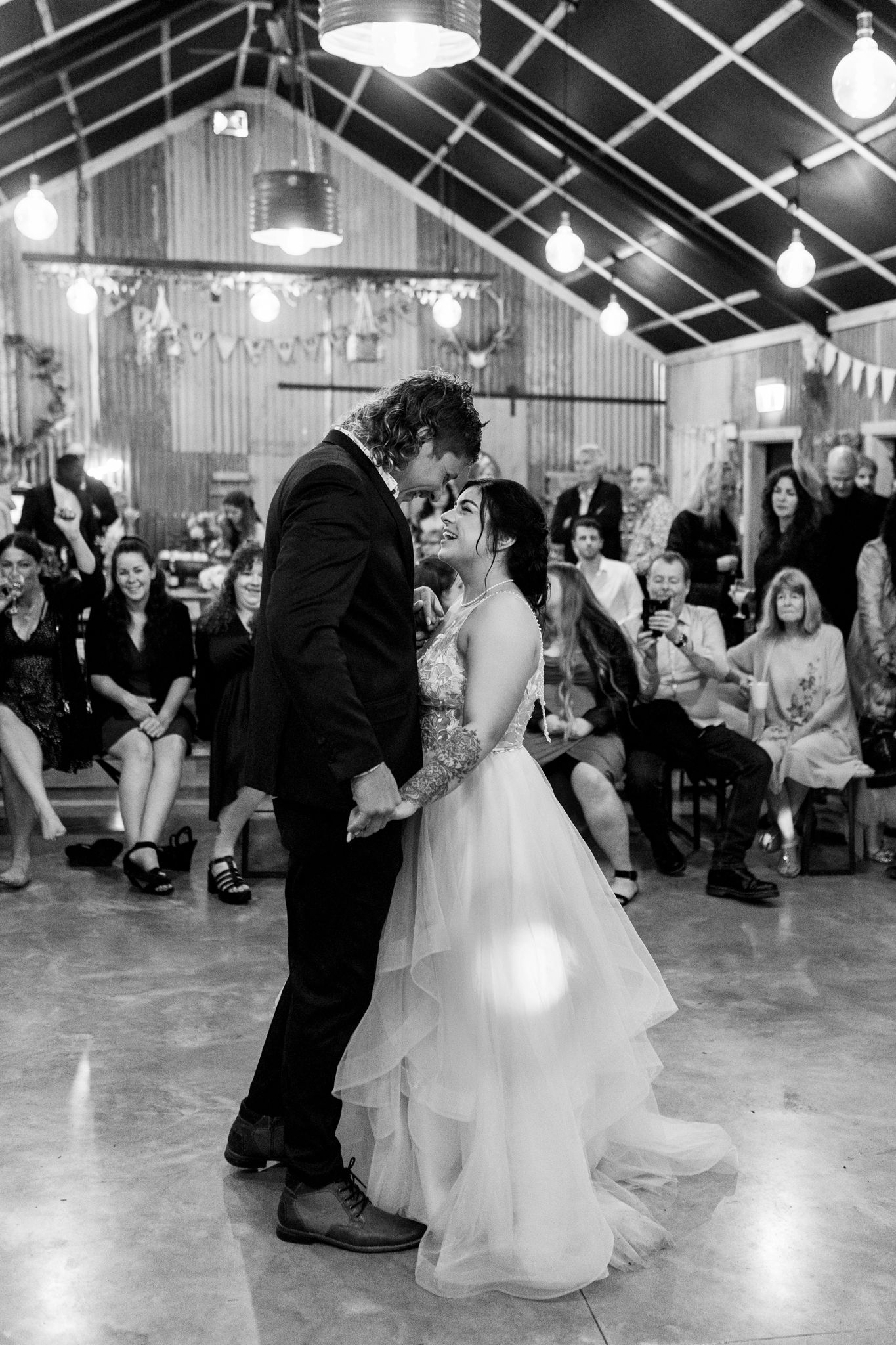 A couple shares their first dance at a rustic barn wedding reception with guests watching from their seats.