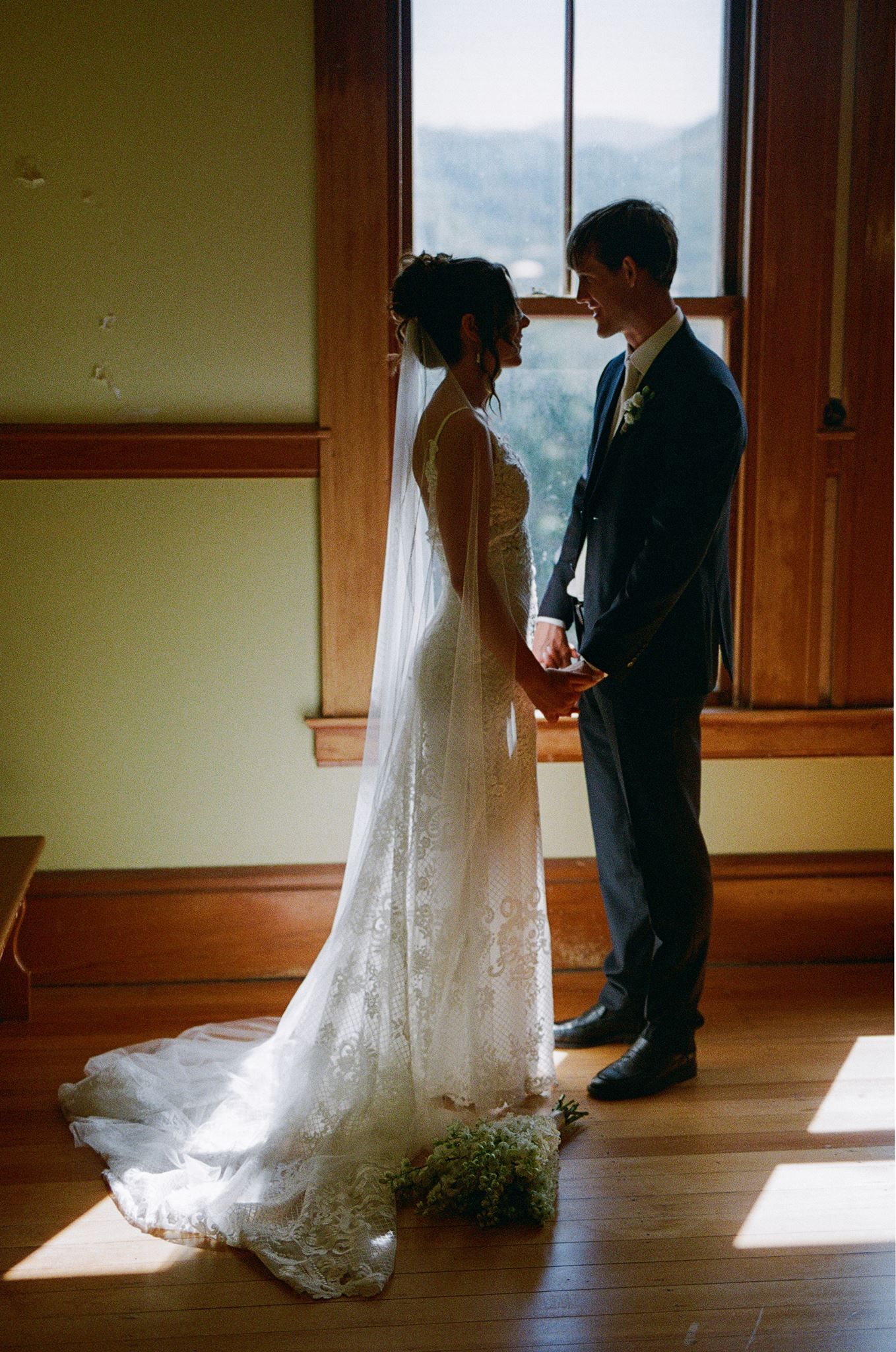 Bride and groom holding hands in golden light by a window, bride wearing lace gown with veil and train.