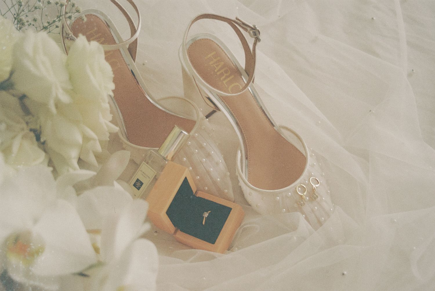 Elegant nude heels and perfume bottle arranged with white flowers and tulle fabric in a romantic flat lay composition.