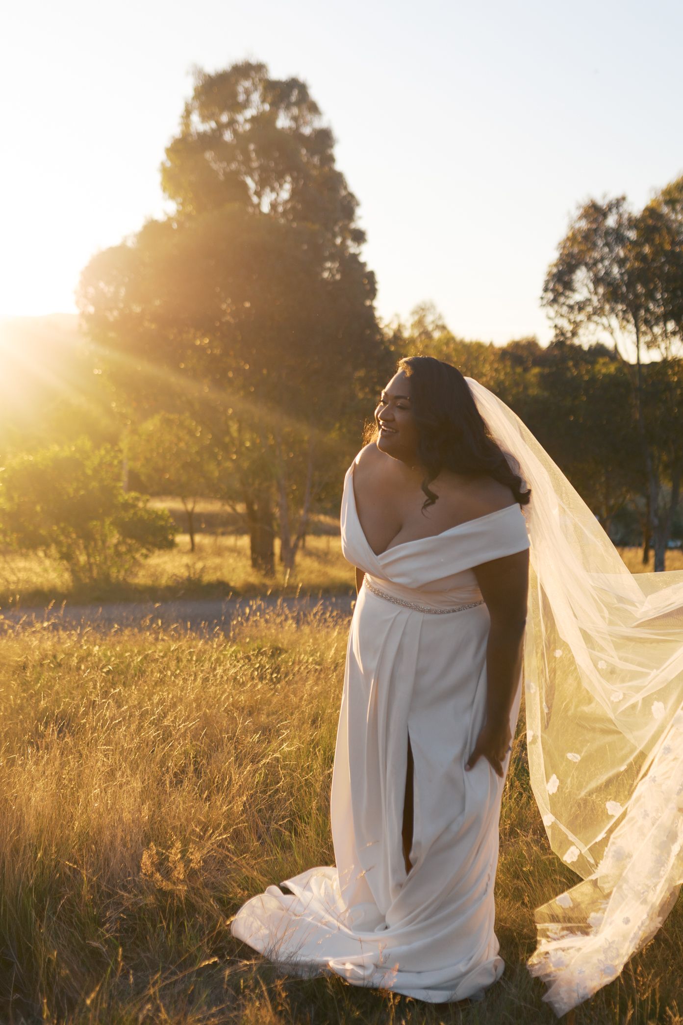 A bride in a flowing white dress stands in a golden field at sunset as her veil catches the evening breeze.