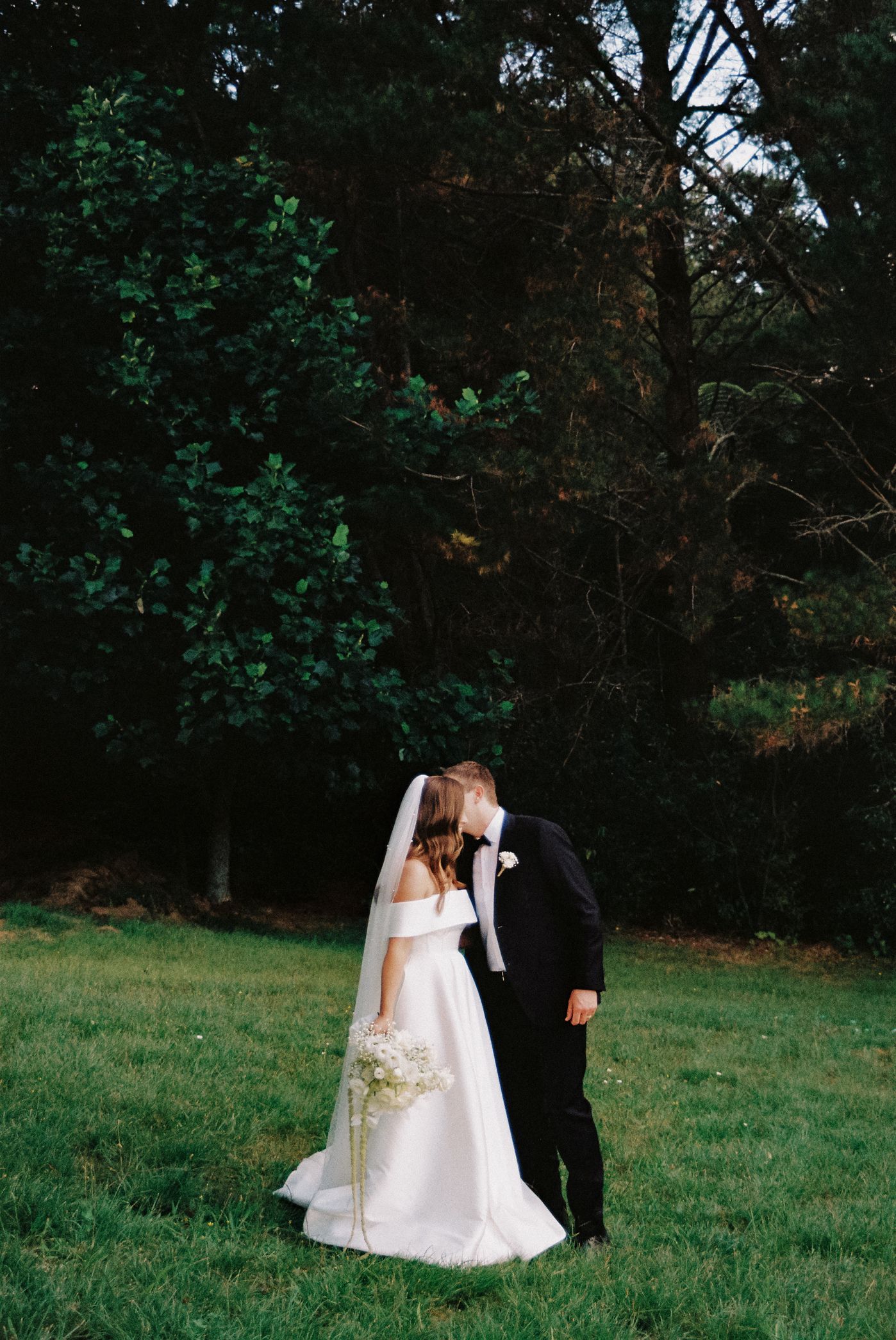A couple in wedding attire shares an intimate moment in a lush green outdoor setting.