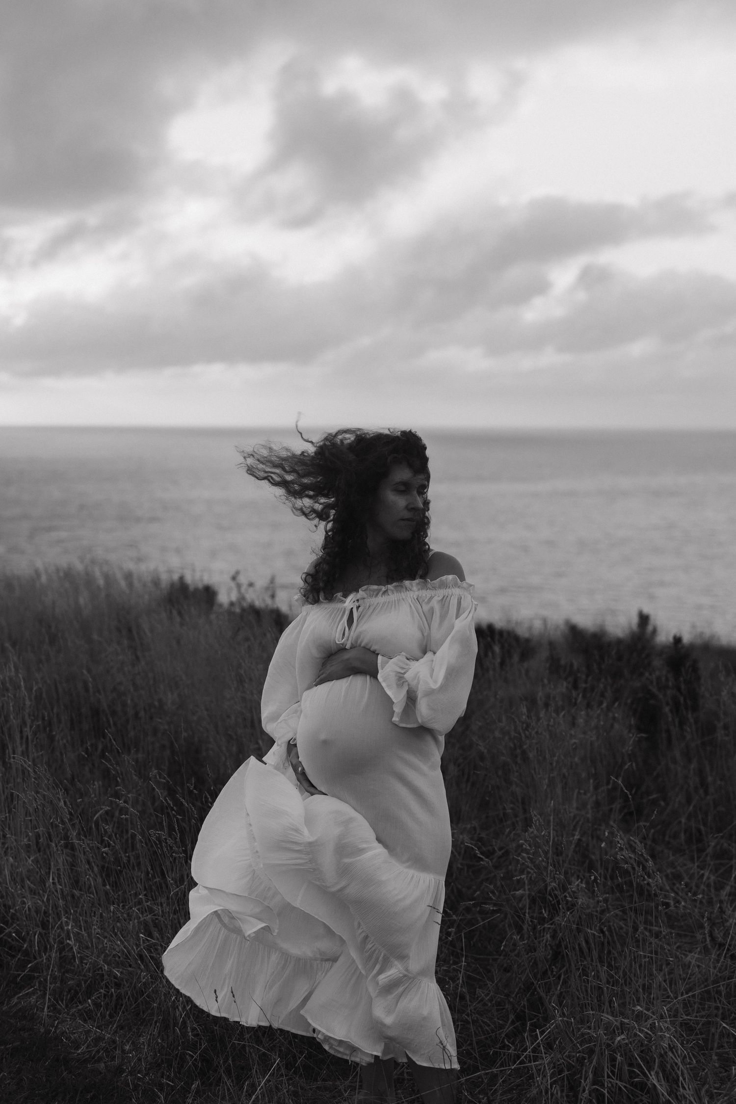 A woman in a flowing white dress stands on a coastal cliff with wind-swept hair against a moody gray sky and ocean.