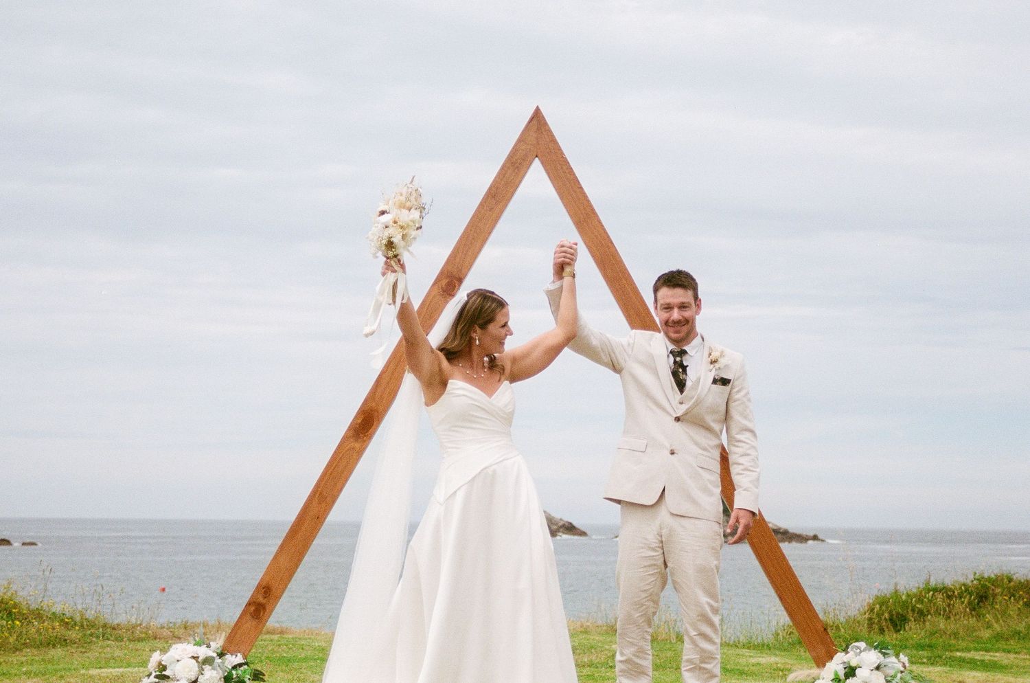 A couple celebrates their wedding ceremony under a wooden triangle arch overlooking the ocean.