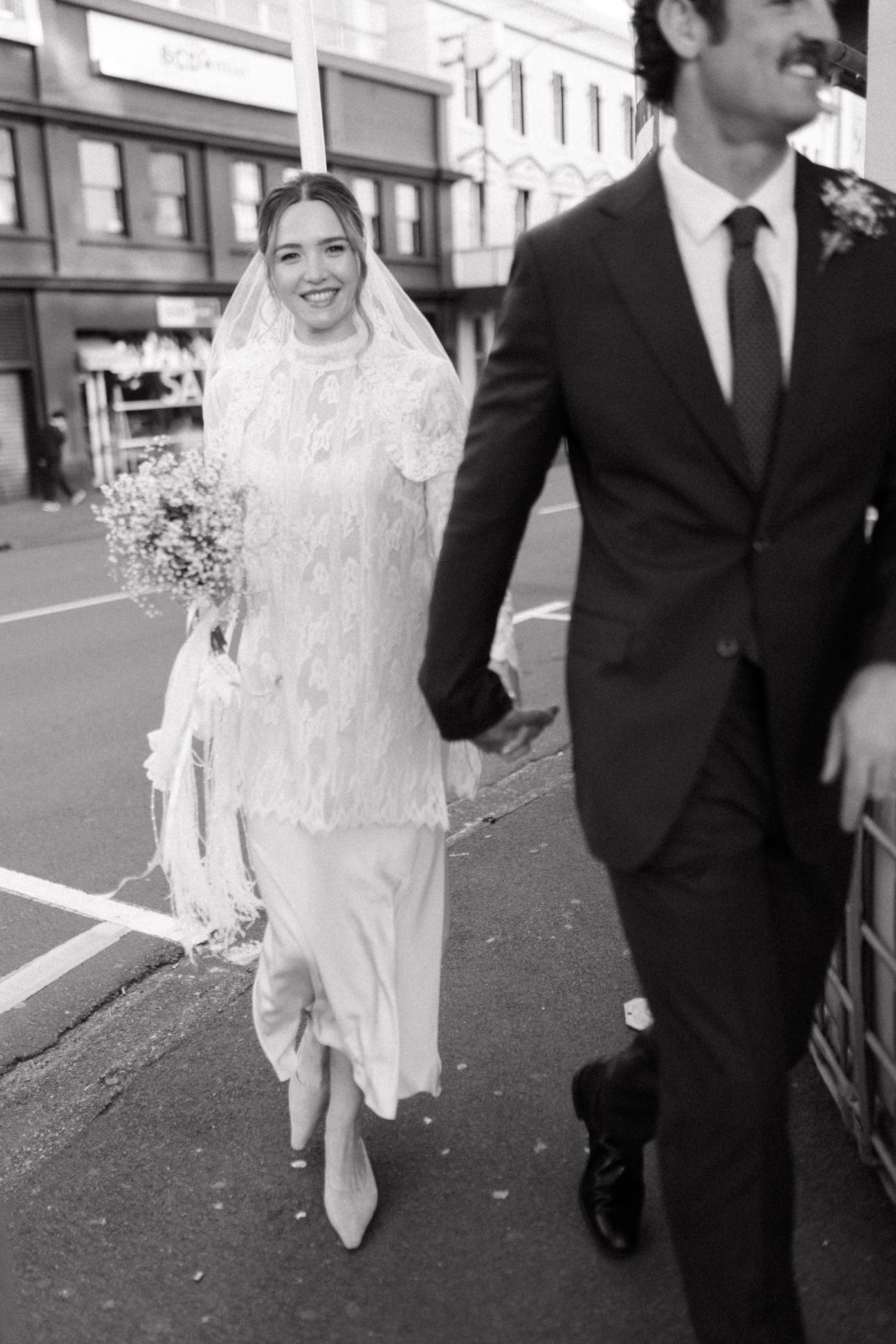 Black and white photo of smiling bride in lace dress and veil holding bouquet, walking with groom in dark suit on city street.