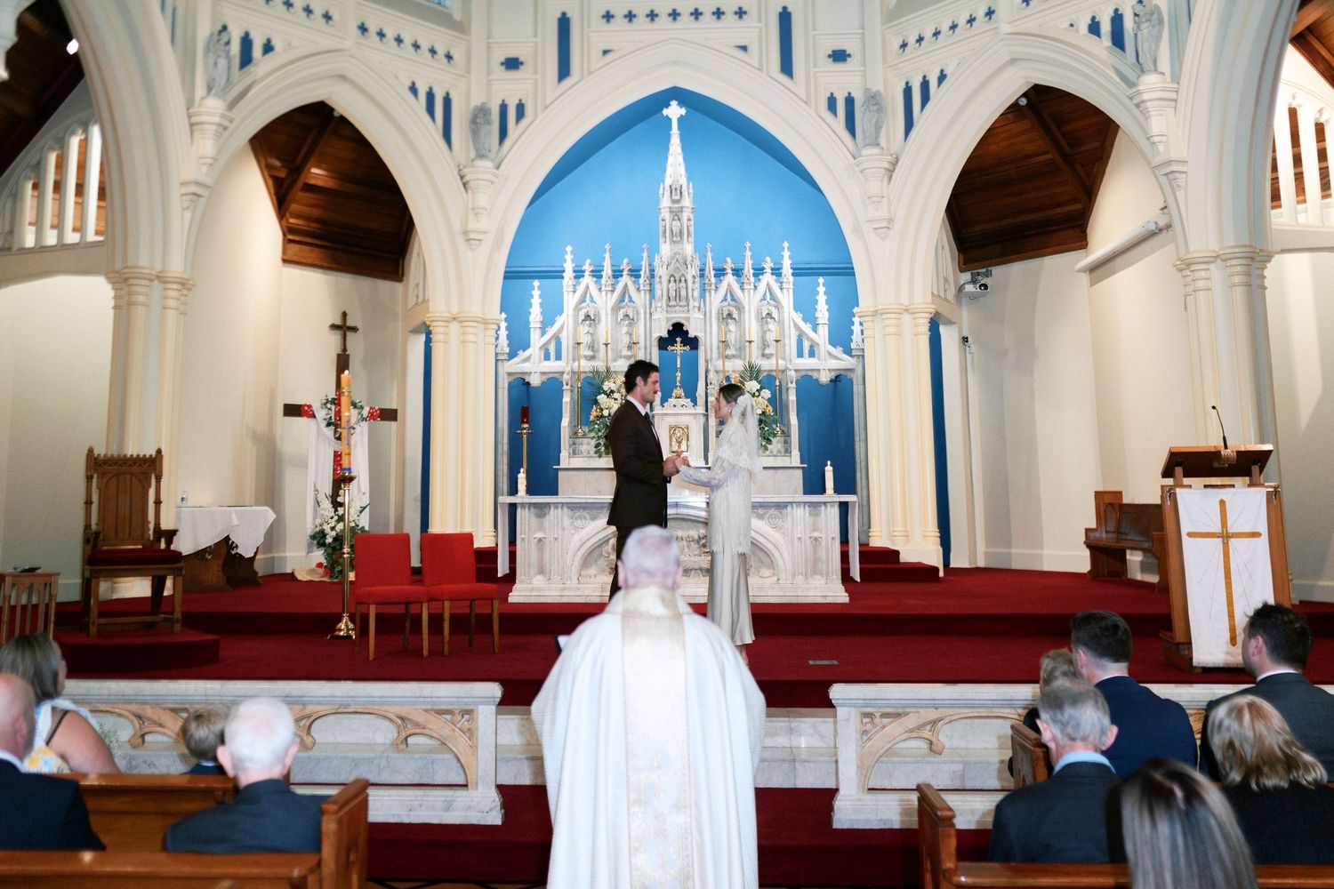 A wedding ceremony at a white church with blue altar, bride and groom at the altar with guests seated in wooden pews.