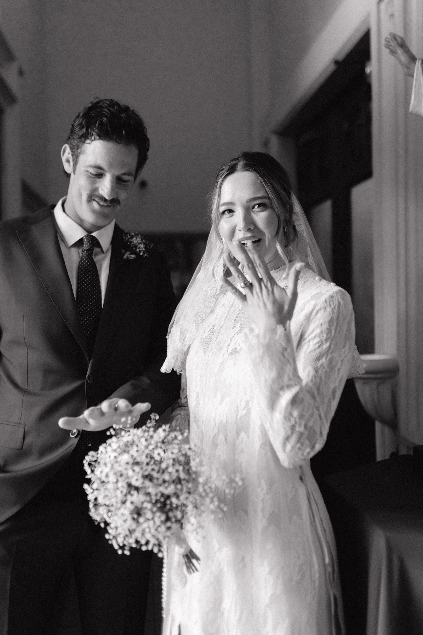 Black and white photo of a smiling bride in lace gown and veil holding bouquet beside groom in suit at wedding ceremony.